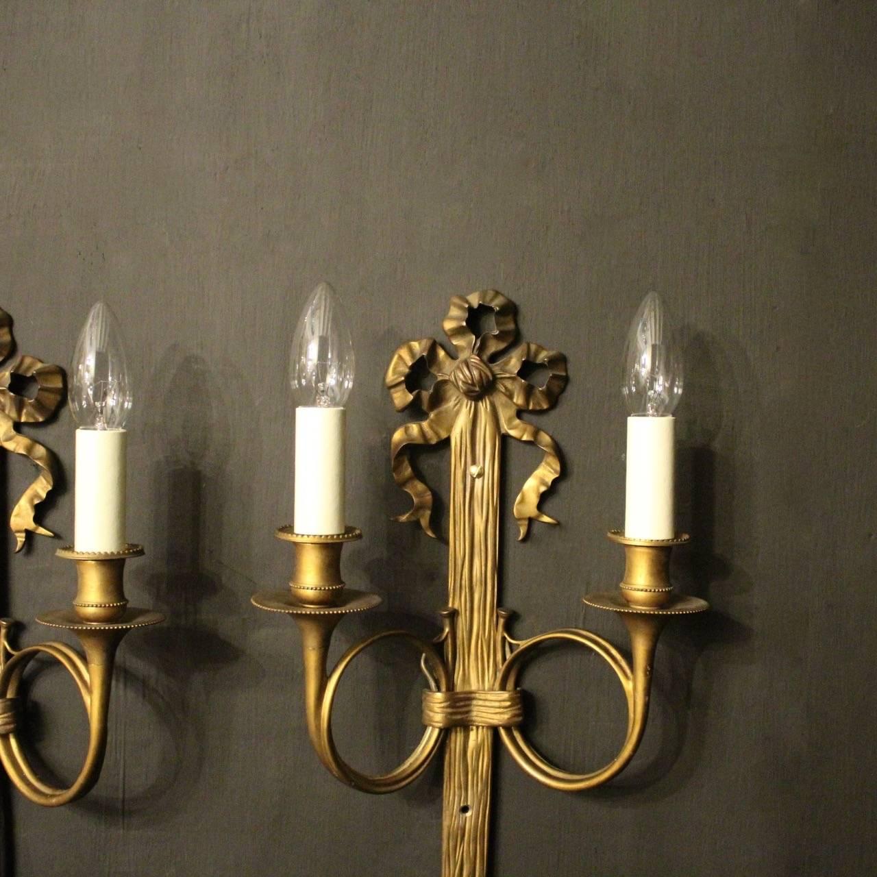 A French pair of gilded bronze twin arm ribbon antique wall sconces, the scrolling arms with trumpet bobeche drip pans and bulbous millgrain candle sconces, issuing from a reeded tassel backplate with pierced ribbon finial, good original gilded