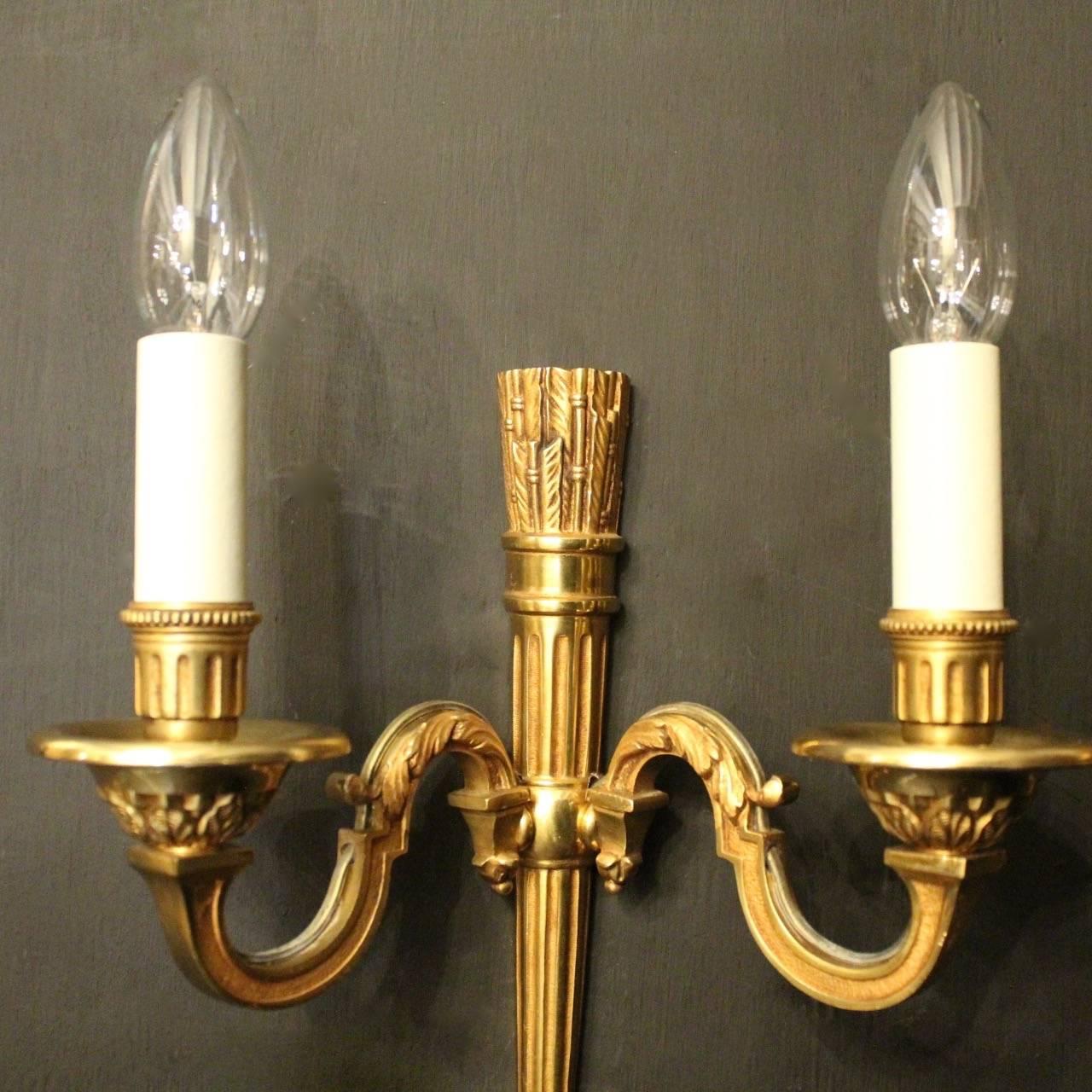 A French pair of gilded bronze twin arm antique wall lights, the ornate leaf square gauge scrolling arms with circular leaf bobeche drip pans with reeded and millgrain candle sconces, issuing from a reeded tapering sheath backplate with arrow head