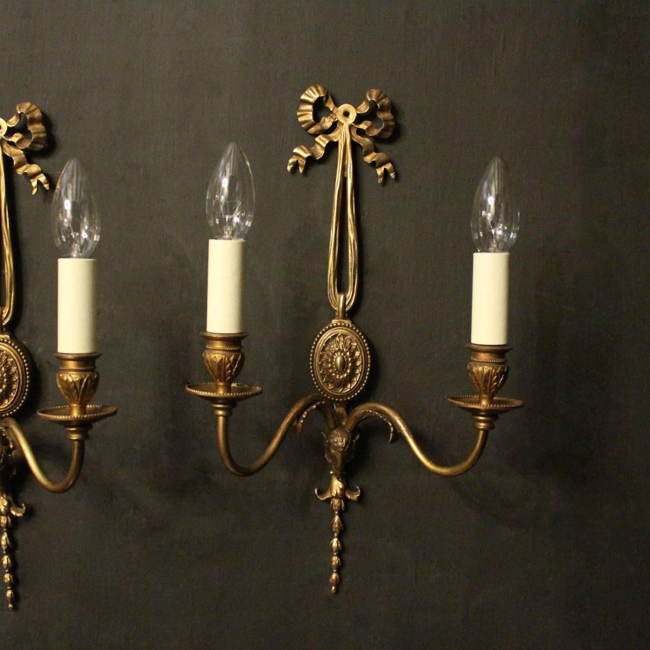 A French pair of gilded bronze twin branch ram headed antique wall lights, the scrolling arms with circular millgrain bobeche drip pans and leaf bulbous candle sconces issuing from an ornate oval central backplate with pierced ribbon finial set