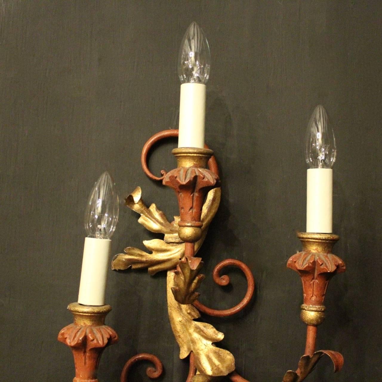 A decorative Italian Florentine carved giltwood and polychrome triple arm opposing wall lights, the foliated tole ware leaf stepped scrolling arms with wooden bulbous candle sconces, issuing from an ornate pierced Acanthus leaf opposing backplate,