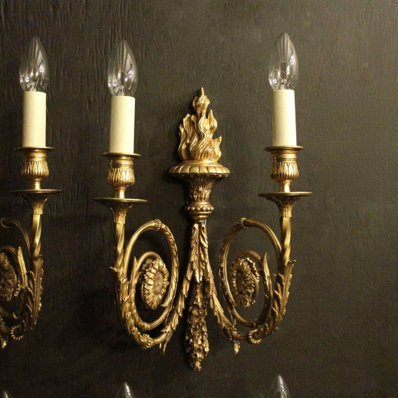 A French set of four gilded bronze twin arm antique wall sconces, the ornate leaf scrolling arms with central flower embellishments and trumpet bobeches drip pans with leaf candle sconces, issuing from a decoratively cast tapering backplate with a