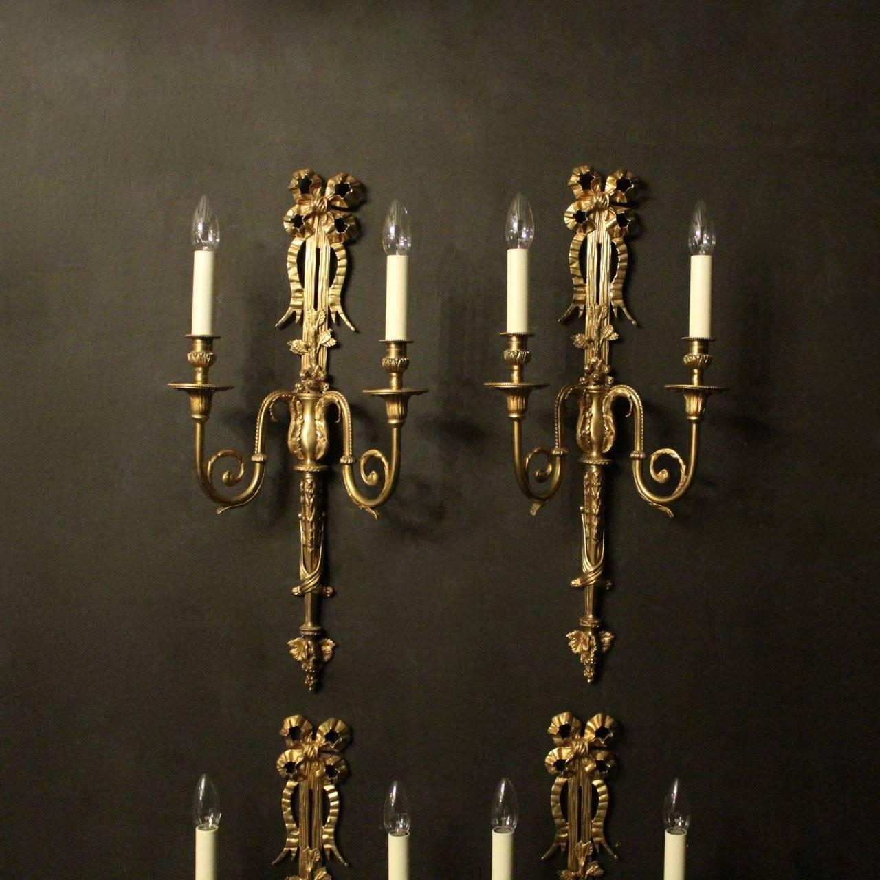 A large set of four French gilded bronze twin-arm antique wall lights, the leaf scrolling arms with reeded trumpet millgrain bobeche drip pans and leaf candle sconces, issuing from a decoratively cast elongated backplate, centring a flower head with