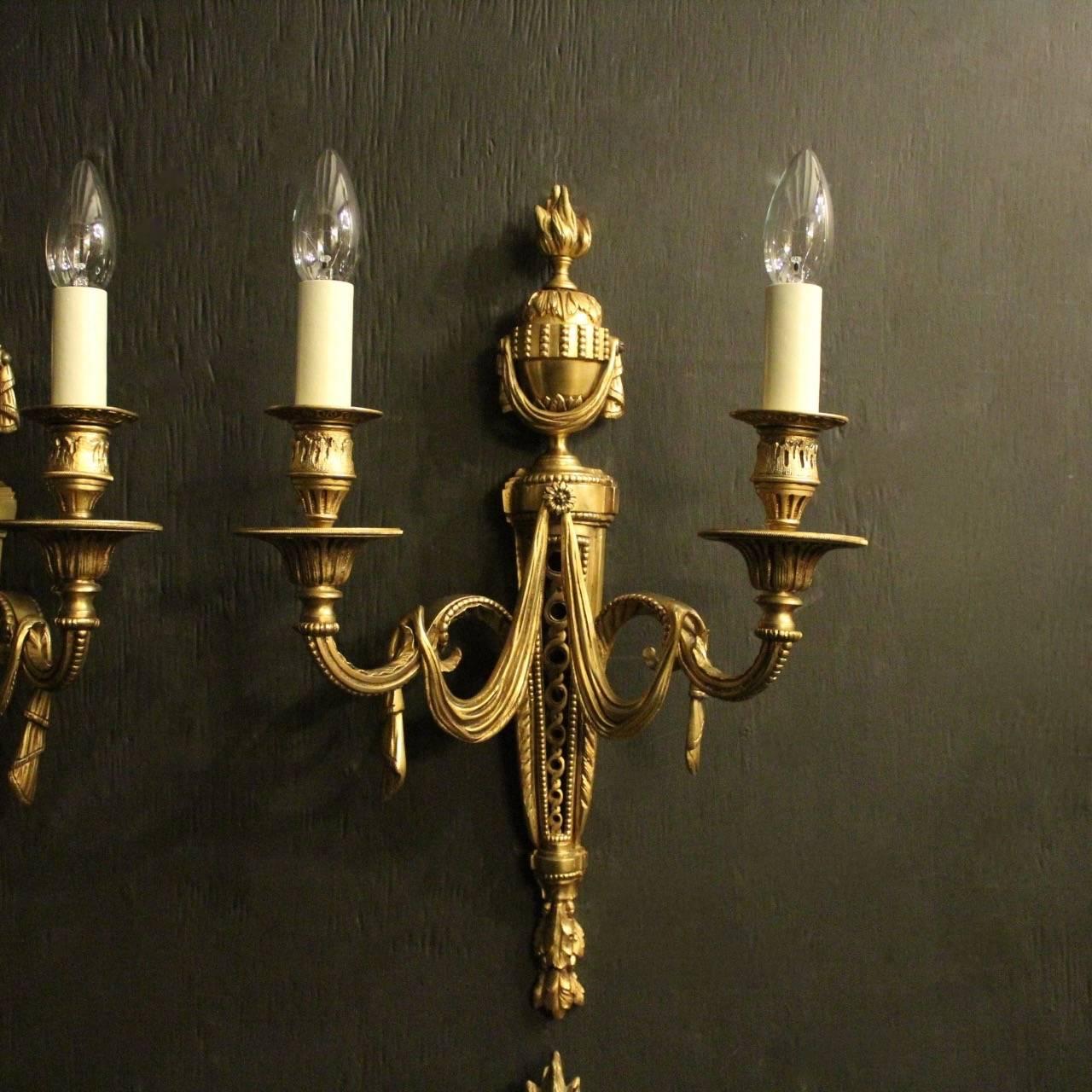 A French set of four gilded bronze twin arm antique wall lights, the leaf scrolling arms with reeded trumpet bobeches drip pans and leaf candle sconces, issuing from a decoratively cast pierced tapering backplate, centring a swaged drapes over both