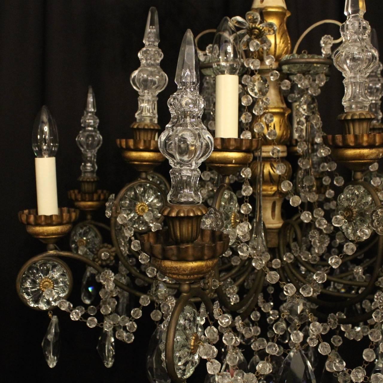 A decorative Italian giltwood, oxidized cast brass and crystal eight light double tiered antique chandelier, the scrolling arms with encased central floral prisms and wooden piecrust bobeche drip pans, issuing from a painted and gilded carved wooden