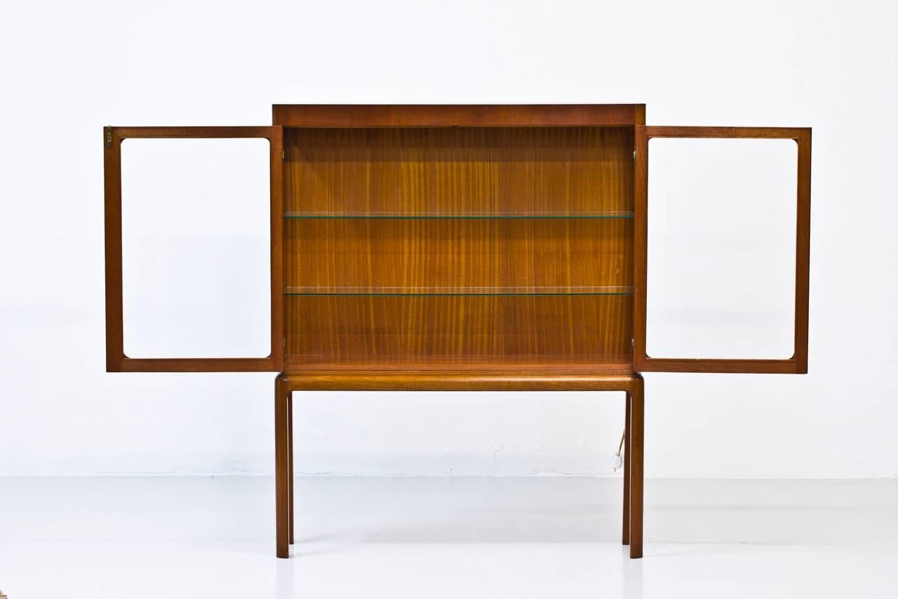 Display cabinet designed by Carl Axel Acking. Produced in Sweden by unknown cabinetmaker in the 1940s. Solid mahogany legs, mahogany, brass and glass. With integrated lamp in working condition. Original key.
 