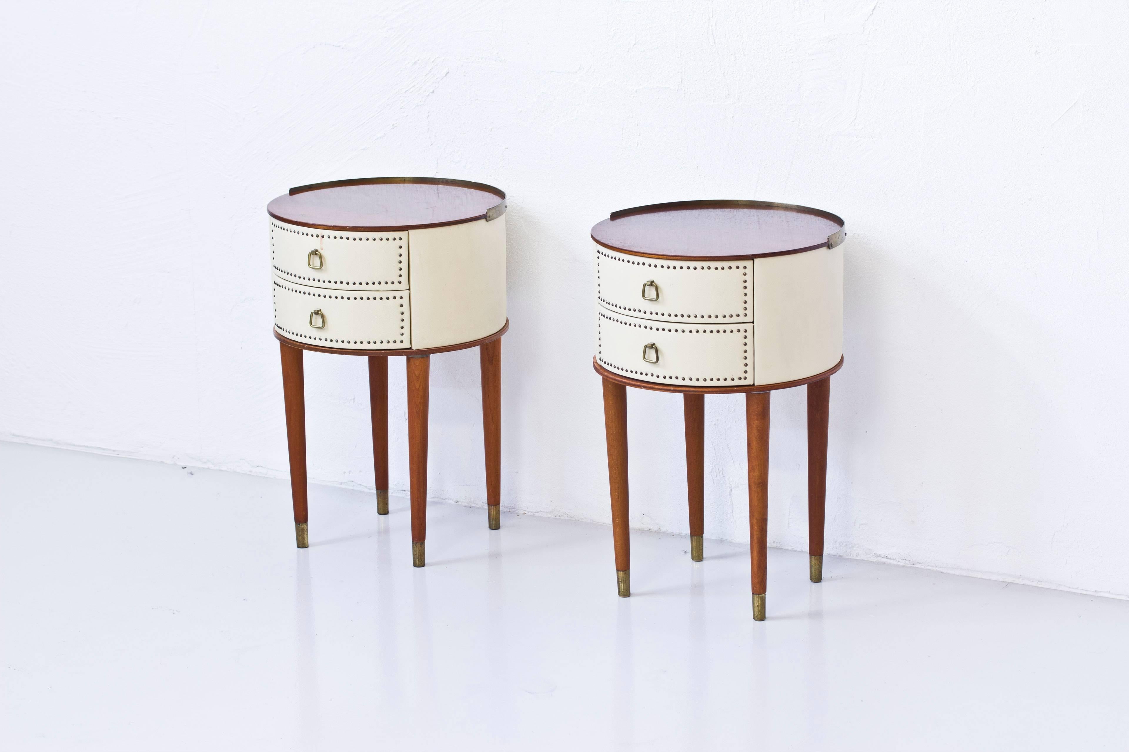 Nightstands designed by Halvdan Pettersson. Produced by his own company in Tibro Sweden during the 1950s. Round wooden cabinets with faux leather, mahogany tabletops and beech legs. Rounded brass edging on the tabletop and brass pulls on the drawers