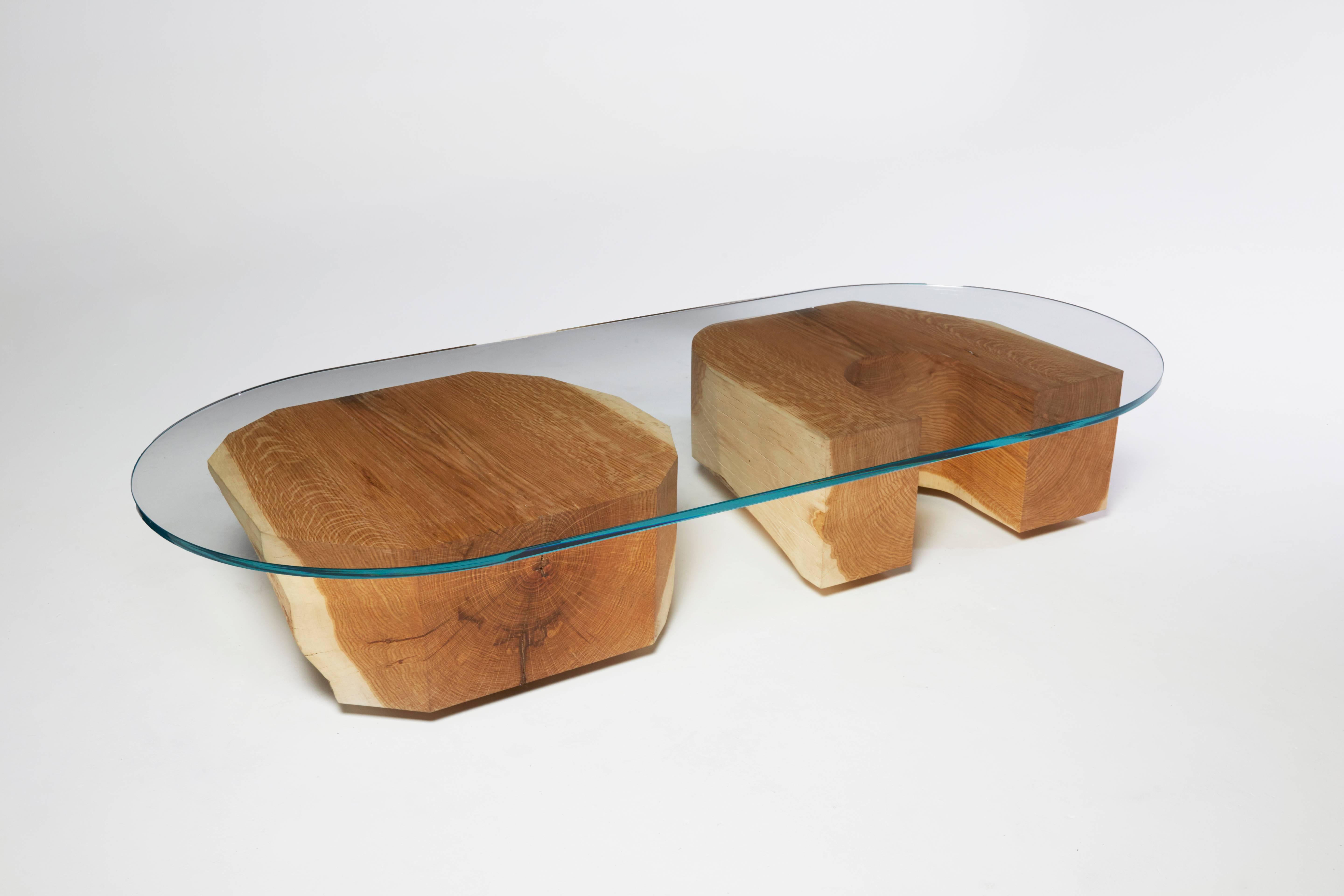 The Tumble Table is a usable sculpture by Noah Spencer made out of two, large, carved pieces of whole white oak and an oval piece of glass. The chunky shapes that hold up the glass tabletop can be placed horizontally on their sides, or vertically