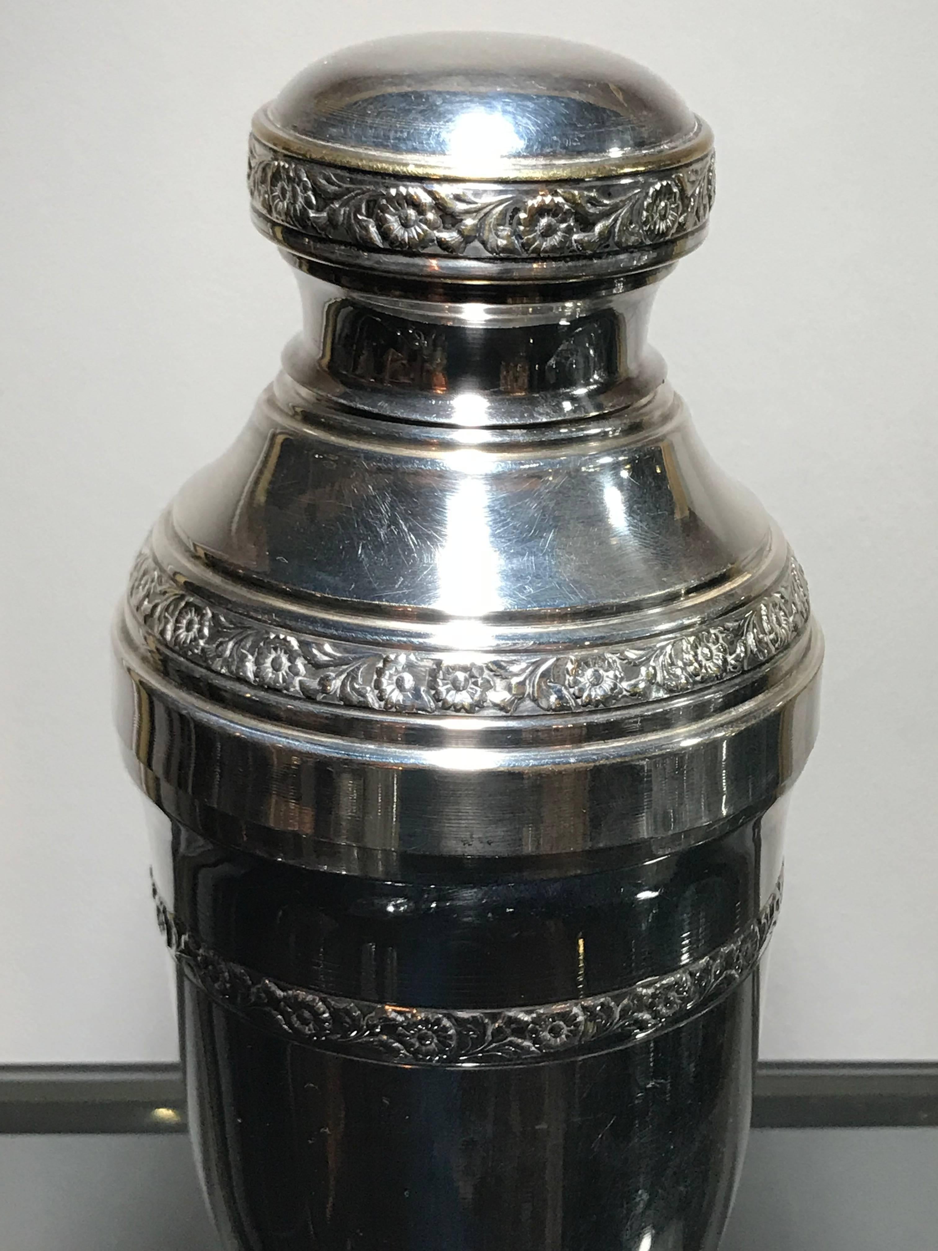 A silver plated cocktail shaker with ornate embossed flowers around the top, shoulder and base. The shaker separates at the shoulder and top, where there is a strainer. The Shaker is in good condition all over with minor marks. Stamped on the base