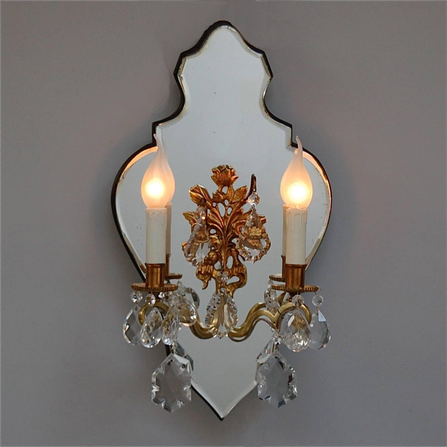 A two-arm candle wall sconce decorated with crystal droplets and mounted on shapely mirror backplate. The centre has a gold colored floral decoration with three hooks from which large crystals are suspended. The mirror has beveled edges and a lovely