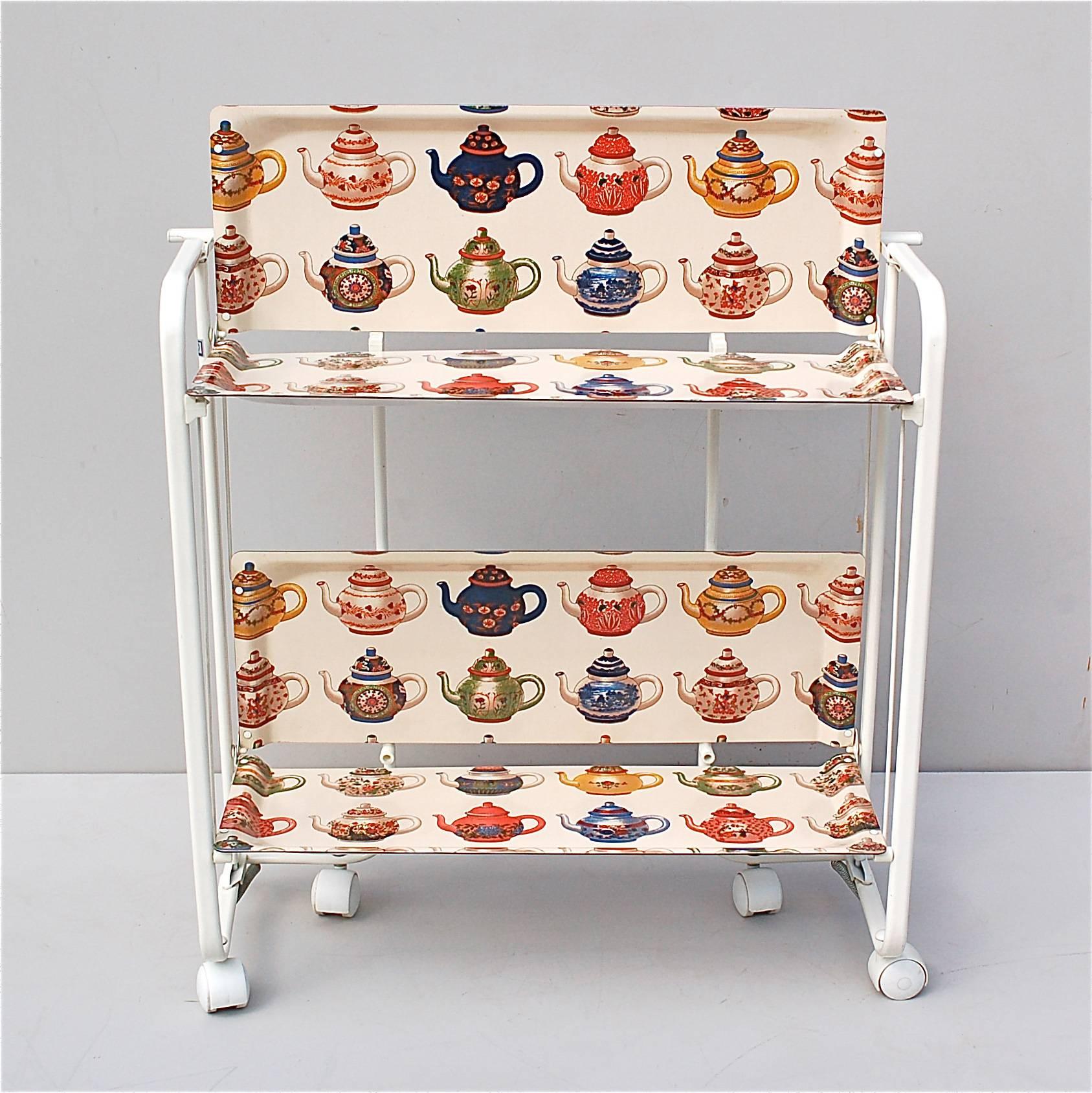 A wonderfully original interpretation of a 1950s drinks or tea trolley on wheels. Made from a white metal frame with two tier formica trays decorated with colorful pattern of tea pots which have a hint of Pierre Frey about them. The trolley can be
