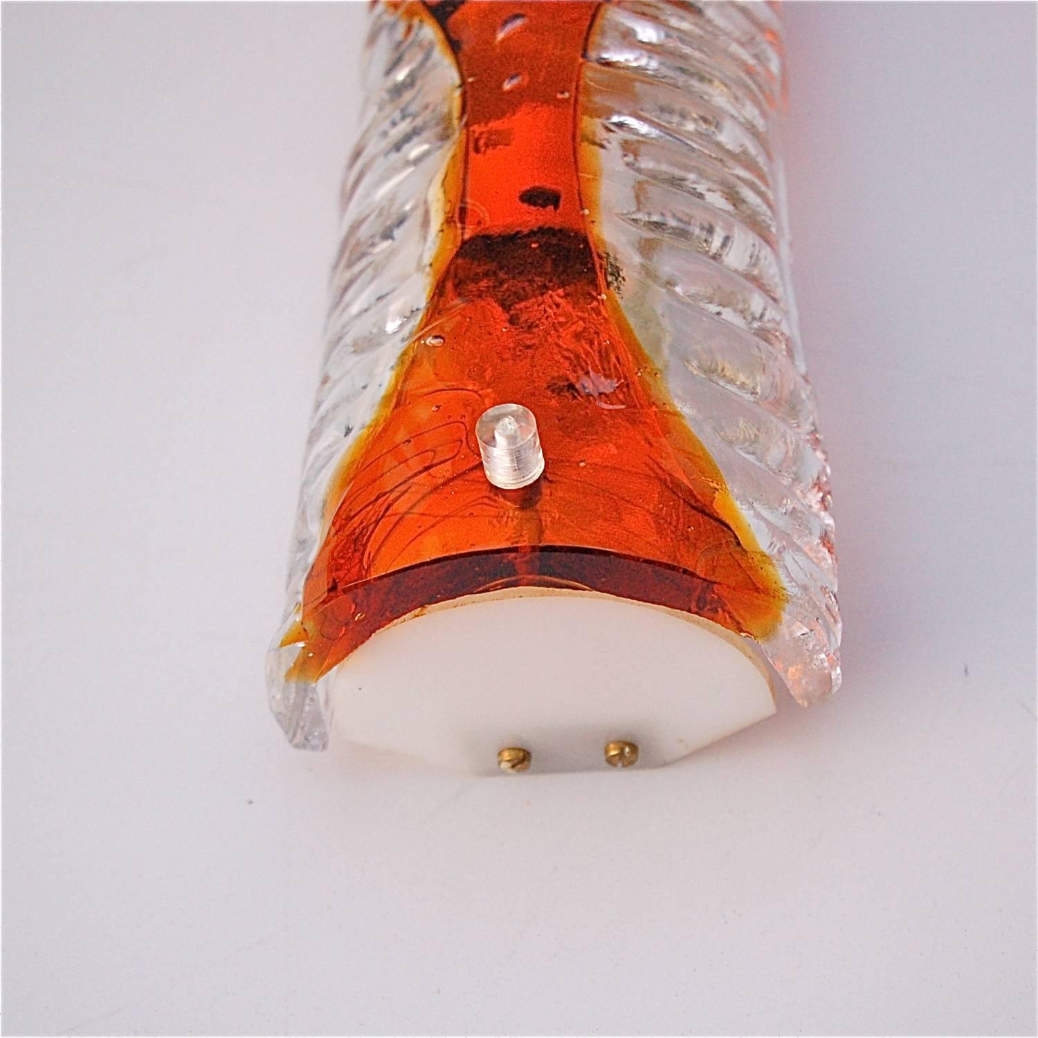 A matching pair of elongated, extended Murano glass wall sconces or wall lights. They are flush mounted and have a curved shape with ribbed effect. They are made of clear, translucent glass with a decorative pattern at the centre in a dark orange.