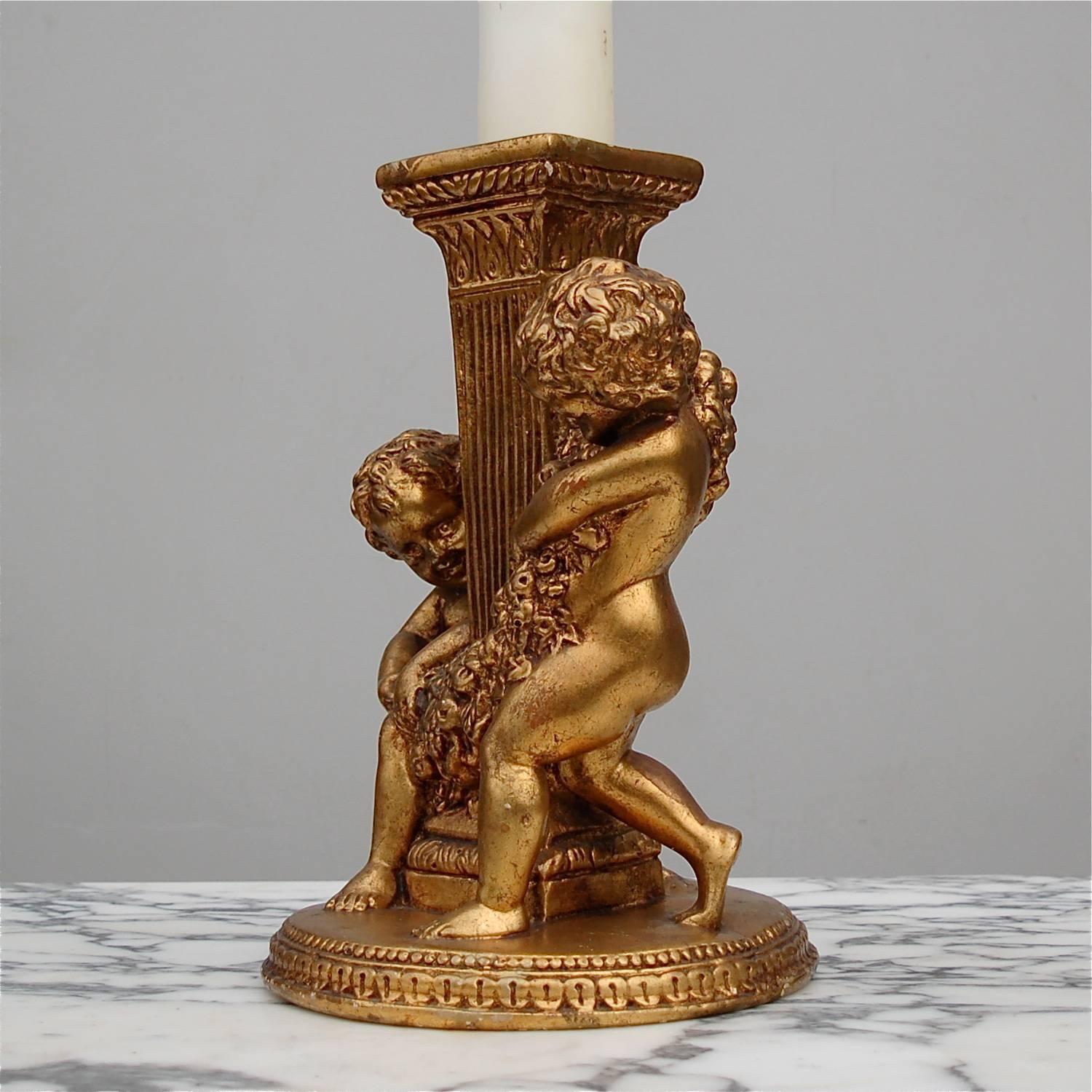 A moulded, gold colored pillar candle holder featuring two adorable little children, resembling cherubs or putti but without the wings. These chubby toddlers are merrily occupied adorning a fluted column with a garland of flowers. This piece has a