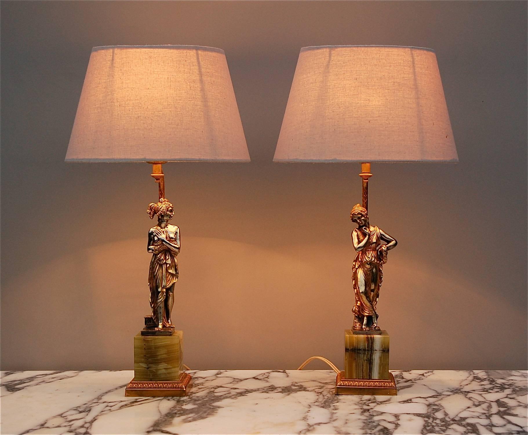 A pair of heavy Italian table lamps with striped onyx base resting on a gold colored brass platform, decorated with a neoclassical motif. A delicately proportioned statue of Roman lady in a draped tunica forms the central focal point of the lamps.