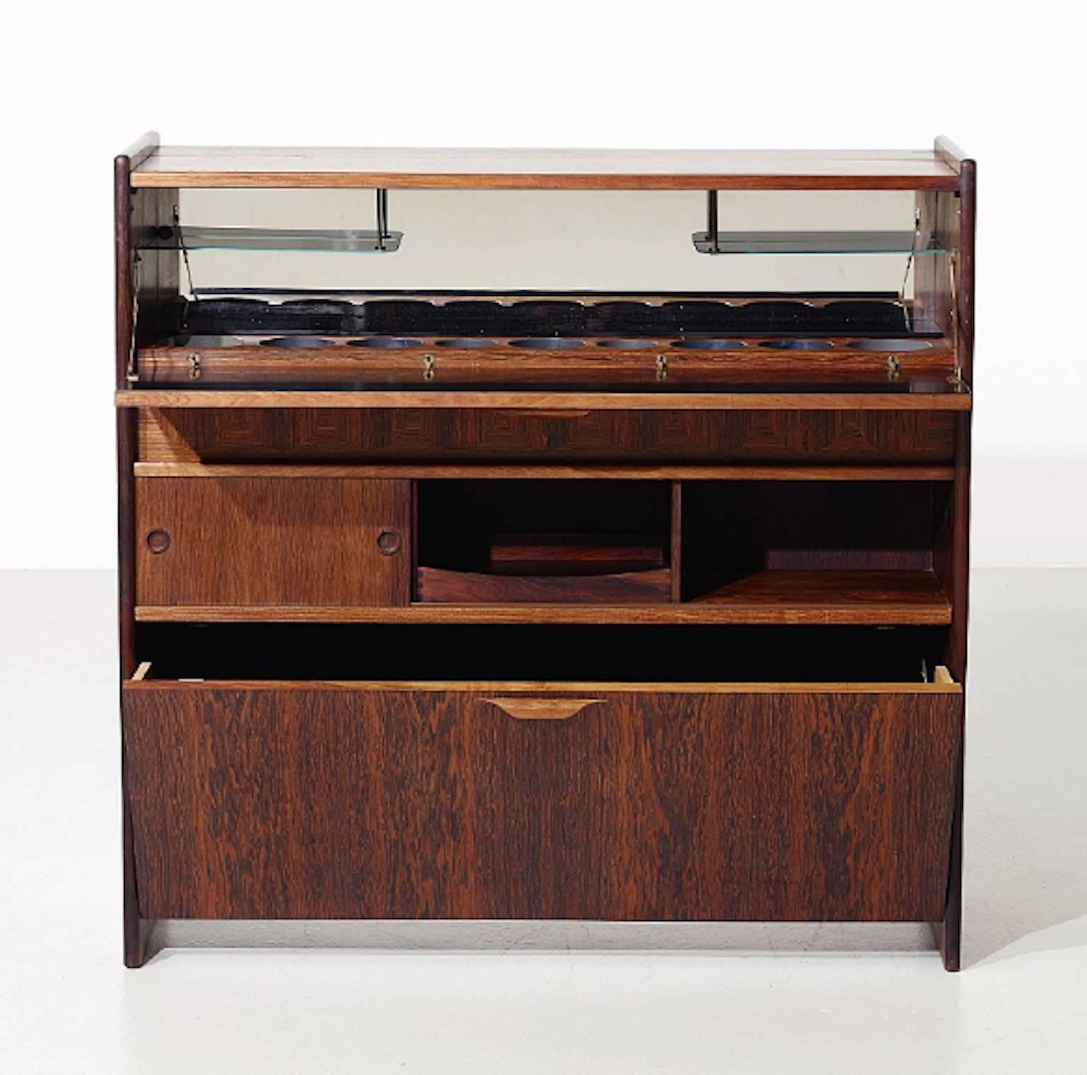 Rosewood bar cabinet designed by Johannes Andersen. The bar is made for serving on one side and sitting on the other side, featuring a mirror, glass shelves, sliding door and space for bottles. Foldable top turns into a bar top; metal rod to rest