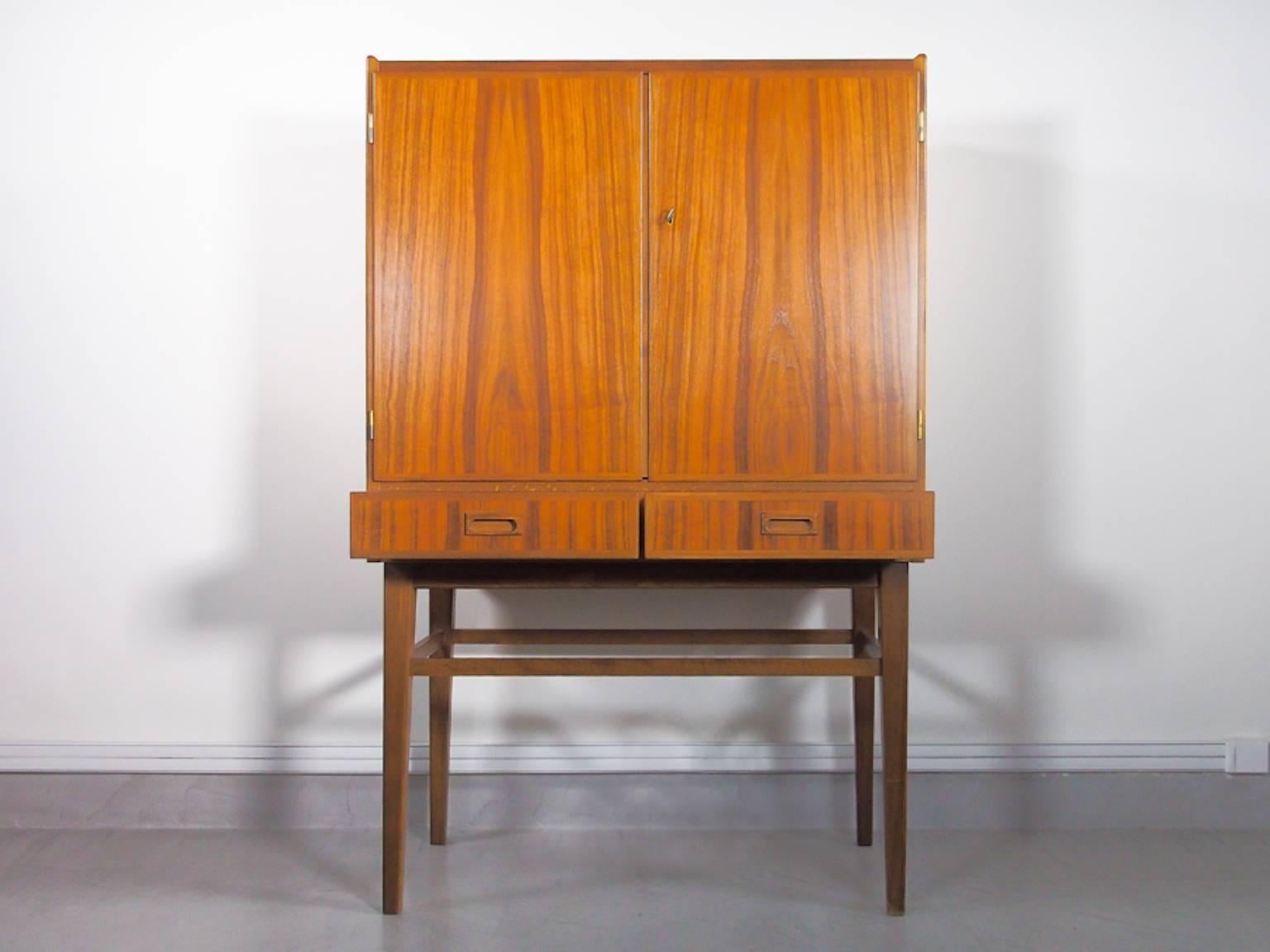 Tall mid-century cocktail cabinet veneered with walnut. Mirrored interior with a glass shelf and a built-in lighting. Underneath, two-drawer and a pull-out table to serve drinks.