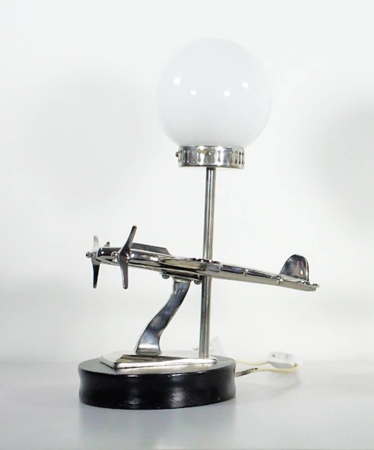 Art Deco style table lamp with an airplane-shaped decoration on a black wooden base. Round shade in white glass, rod and aircraft design in chromed metal.