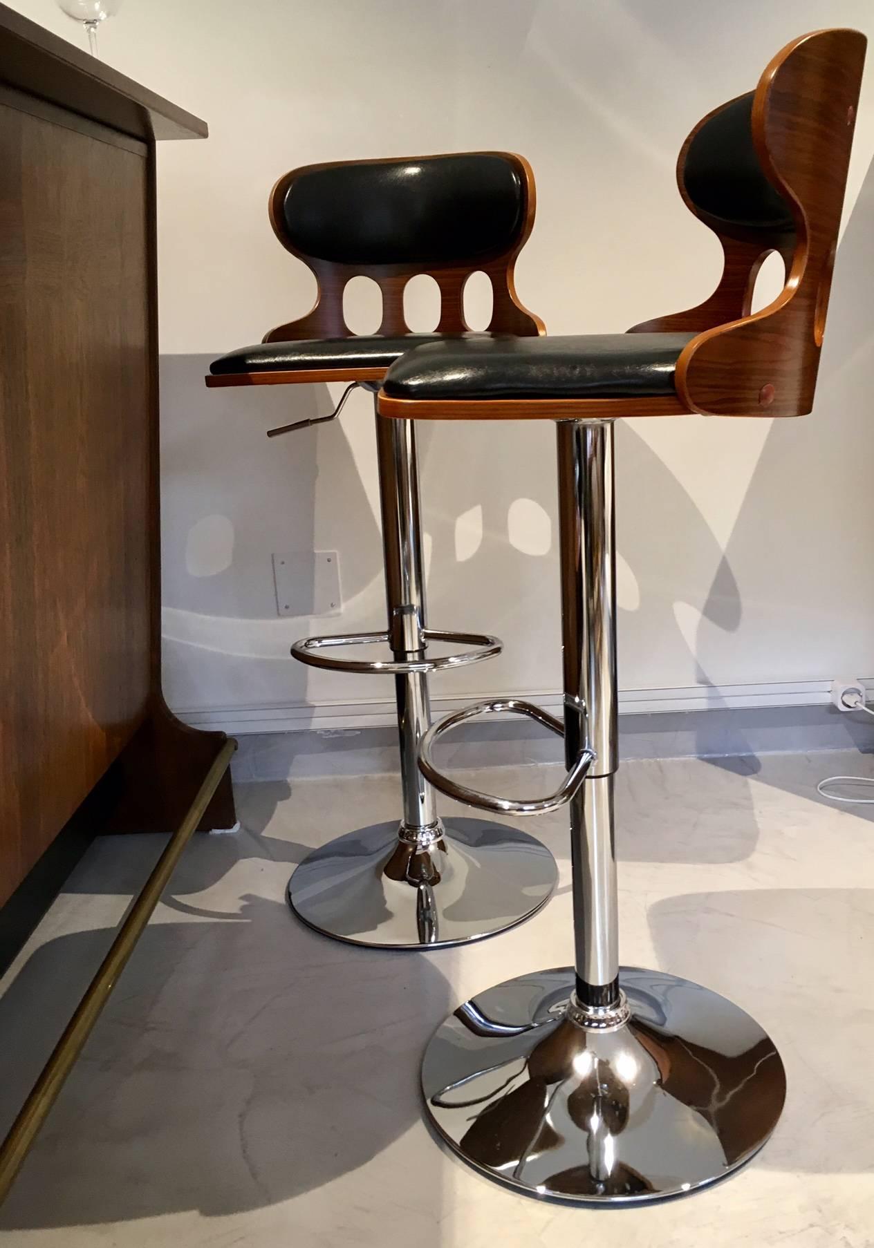 Two high chromed metal bar or kitchen stools with black faux leather seat upholstery and brown wood veneer exterior. Seat increase or decrease height 80/60 cm.