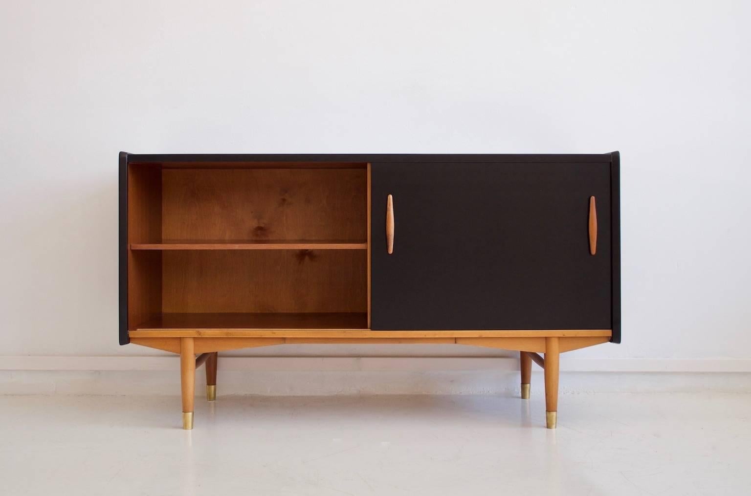 Teak veneer sideboard with black laminate front door and five drawers designed by Hugo Troeds in 1957. Legs in beech with brass finish. Back labeled with Hugo Troeds, Bjärnum.