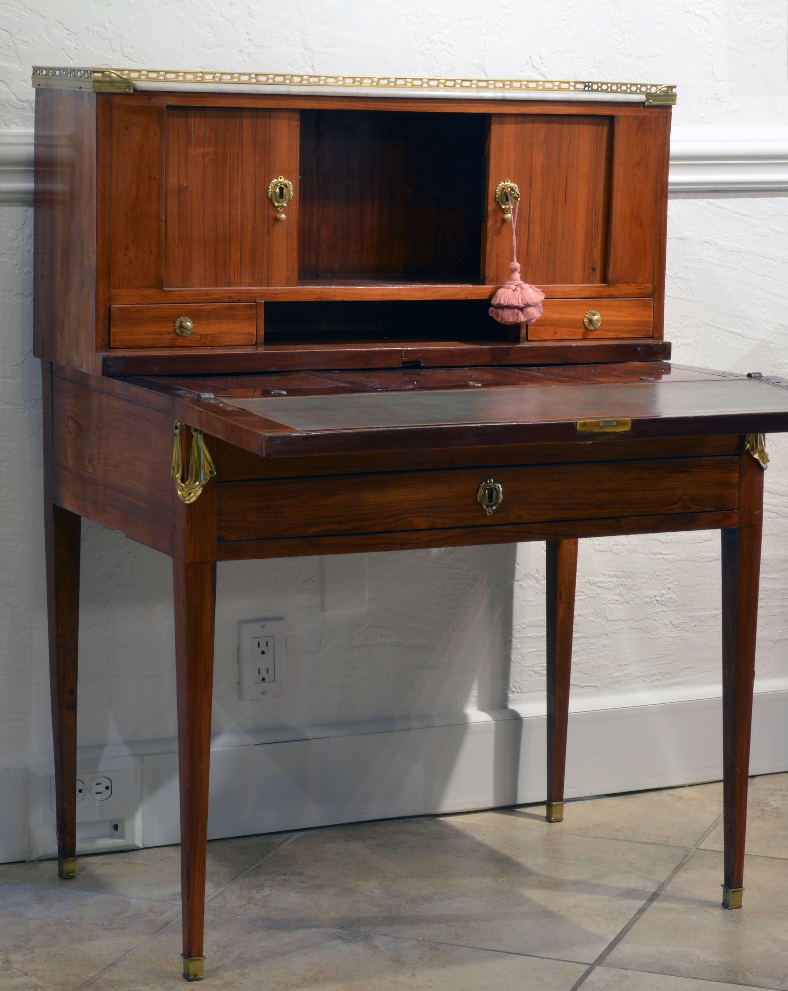 This Louis XVI 'Bonheur du Jour' (ladies writing desk) features an upper part with gallery edged marble-top above two tambour doors enclosing an open compartment under which two small drawers flank an open convenience space. The lower part features