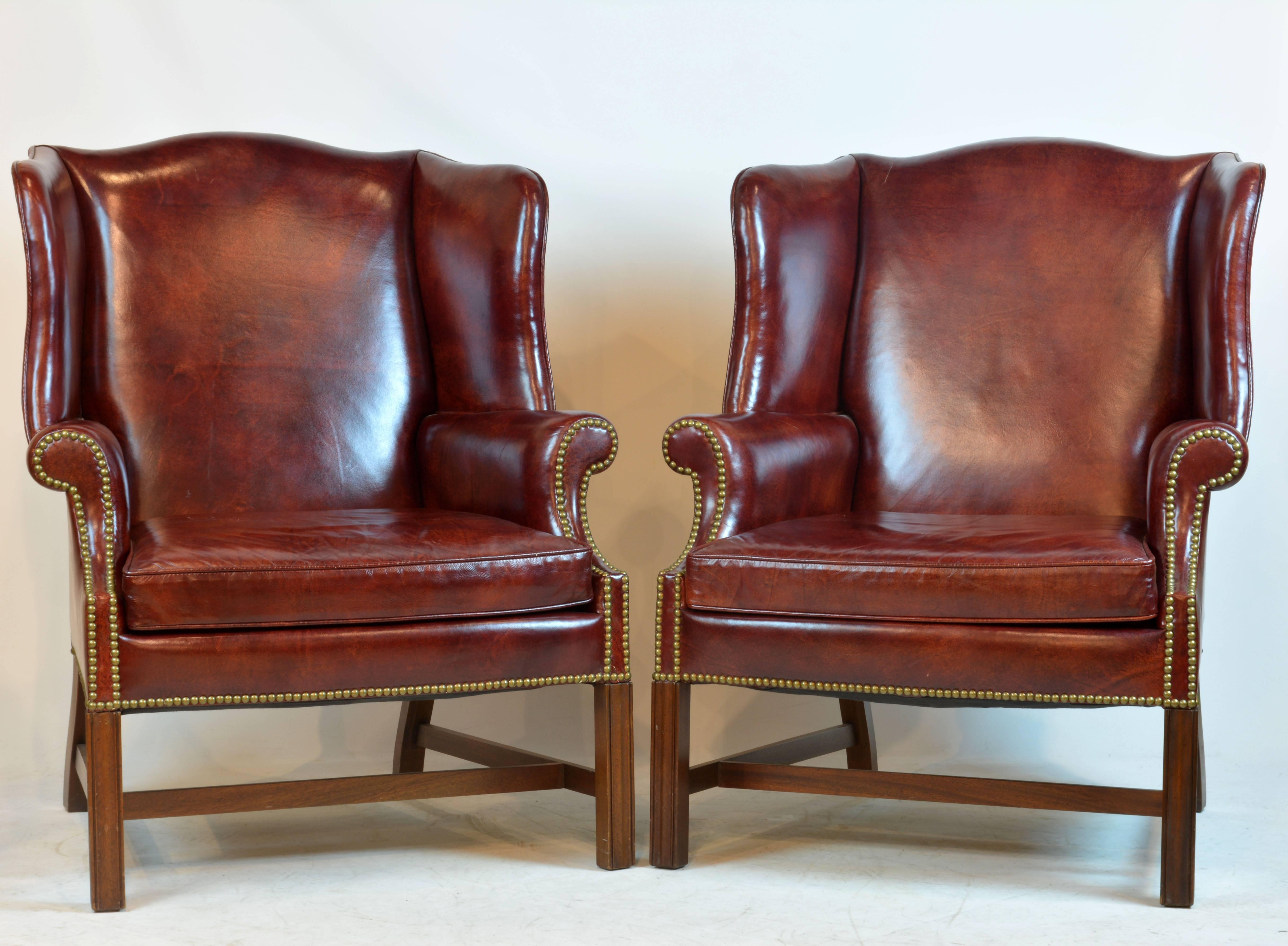 Beautifully accented by traditional brass nailhead trim these fine Georgian style burgundy brown leather covered chairs feature large scrolled wings above a loose cushion seat supported by four square chamfered legs joined by stretchers.