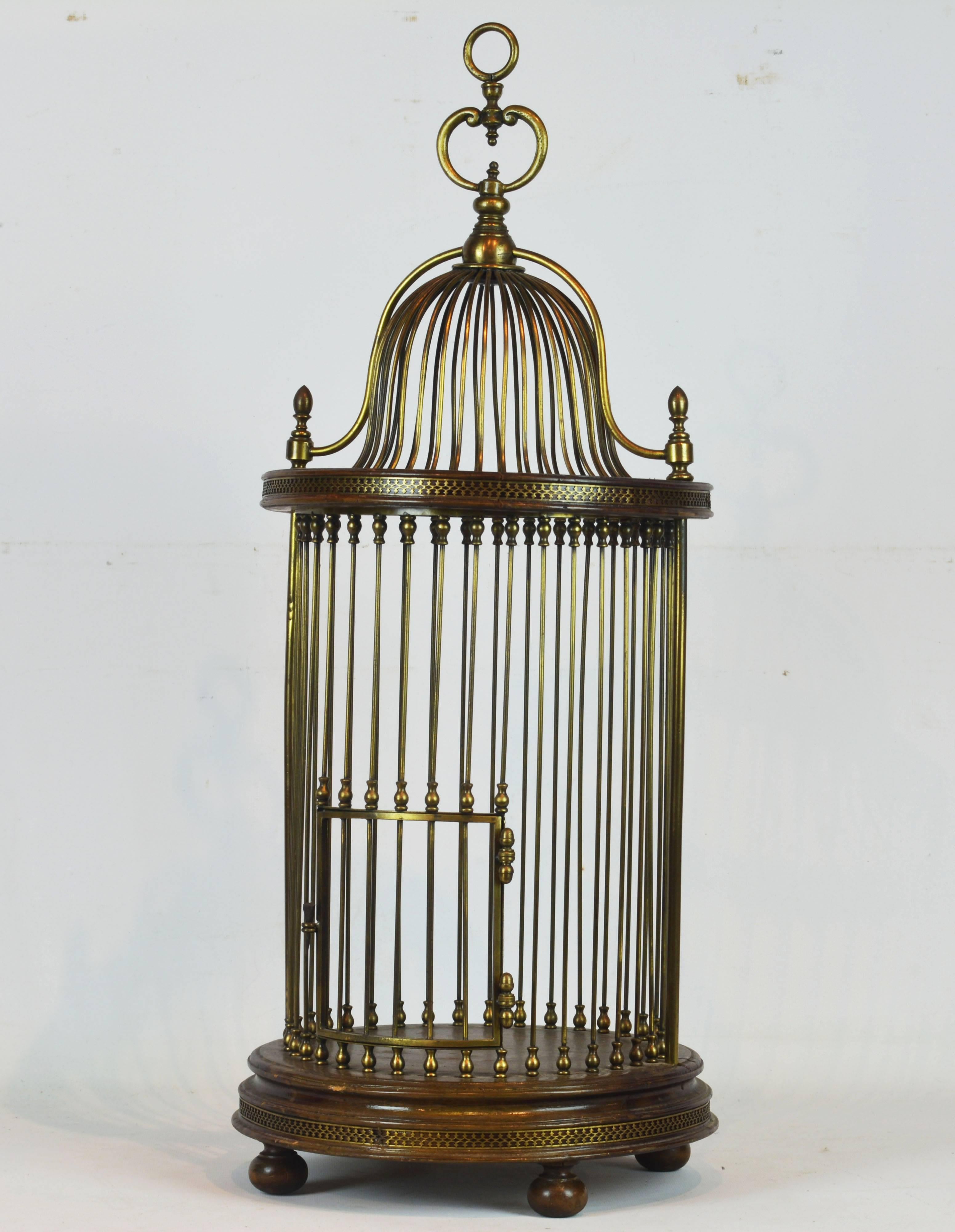 Raised on four bun feet the wooden base platform, accented by reticulated brass bands, supports the circular cage with hinged door and a wooden pediment likewise embellished and surmounted by the oriental style cupola.
