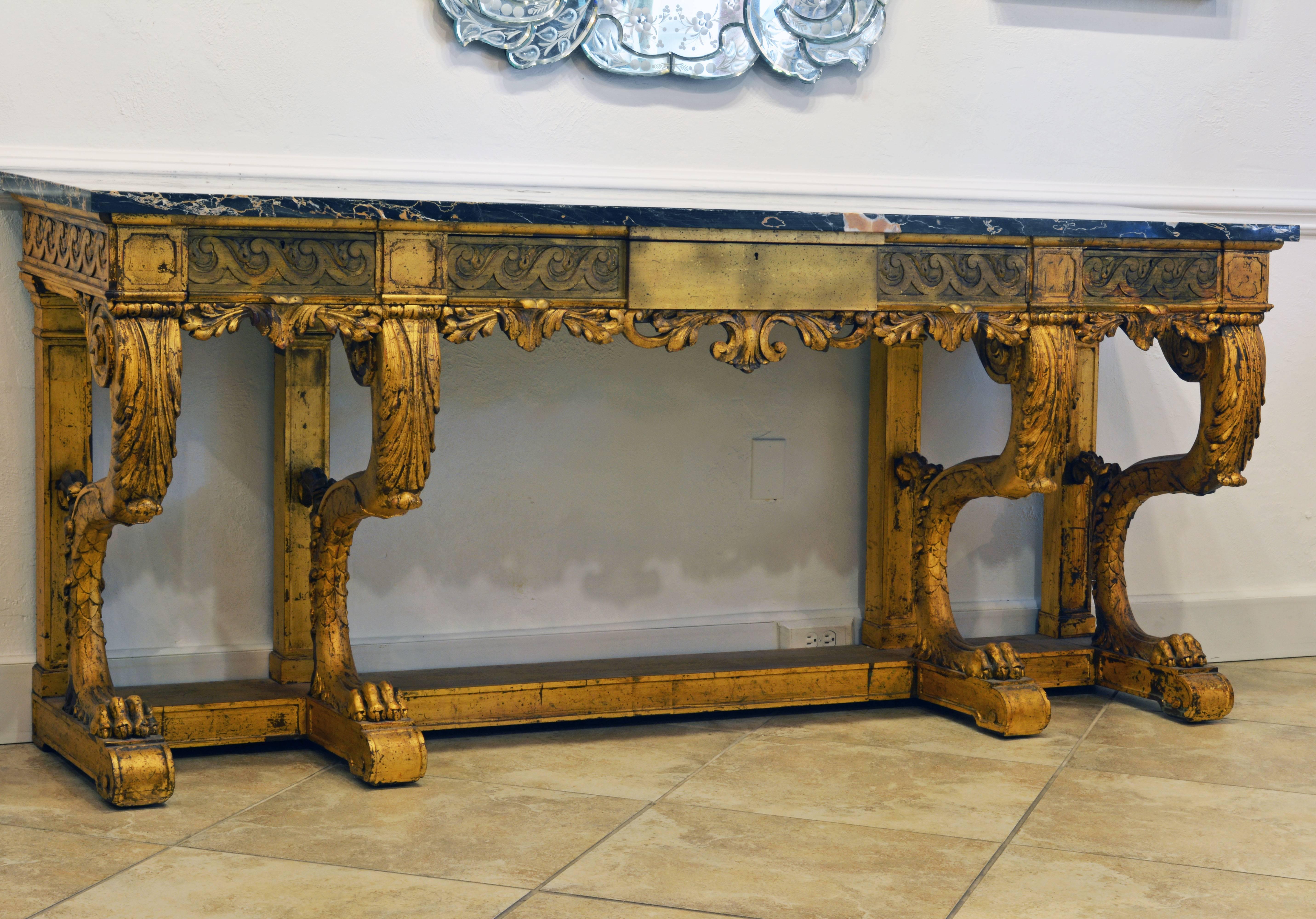 Made likely in the 1920s this palatial carved giltwood and marble top console table features a grey, white and ocher veined marble-top above a Vitruvian scroll carved frieze with a central long drawer flanked by two shorter drawers above an open