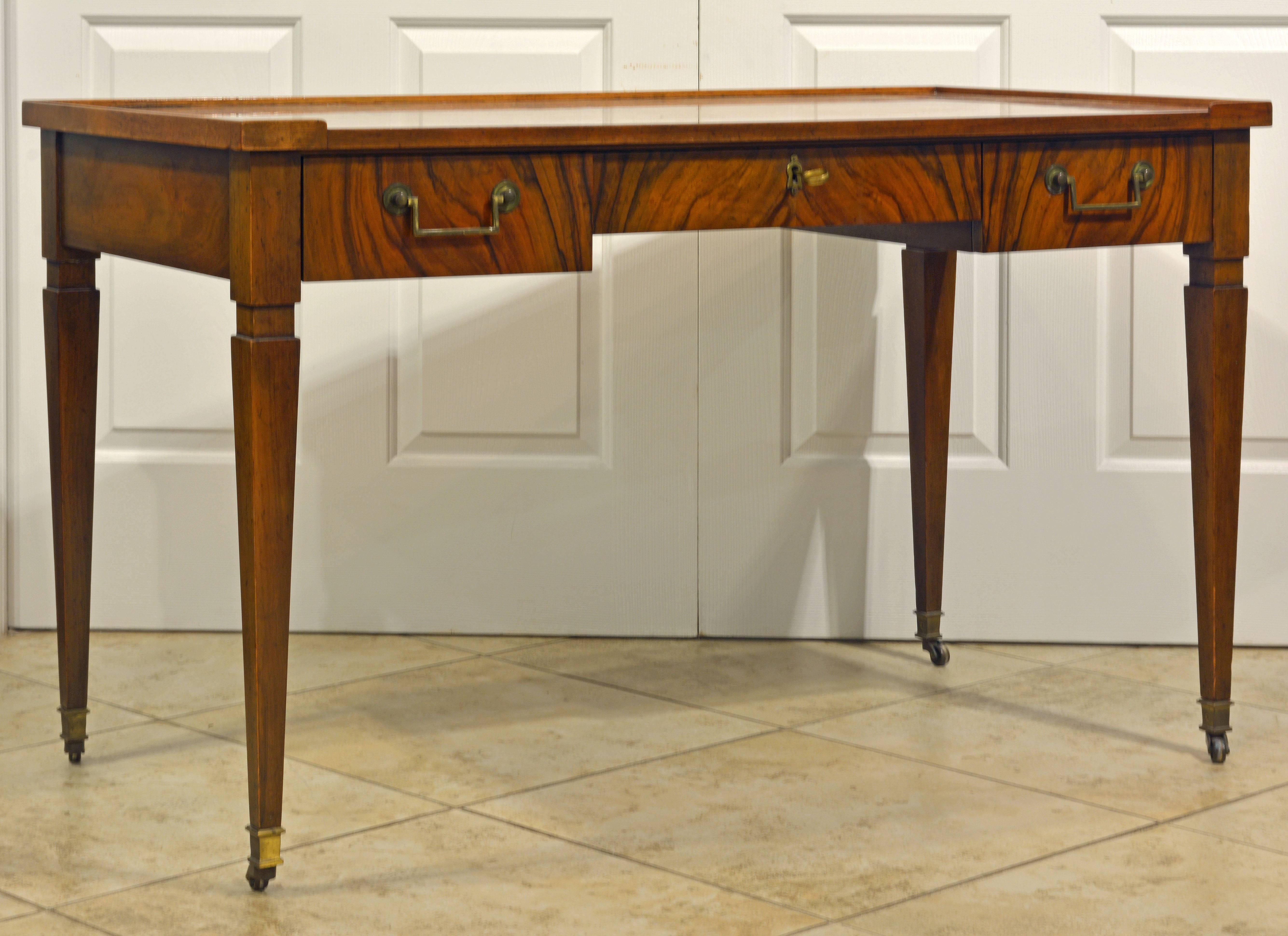 This Classic desk of versatile dimensions features a beautifully figured walnut writing surface surrounded by a gallery lip on three sides above three frieze drawers with solid brass handles resting on square tapering legs and brass casters. In