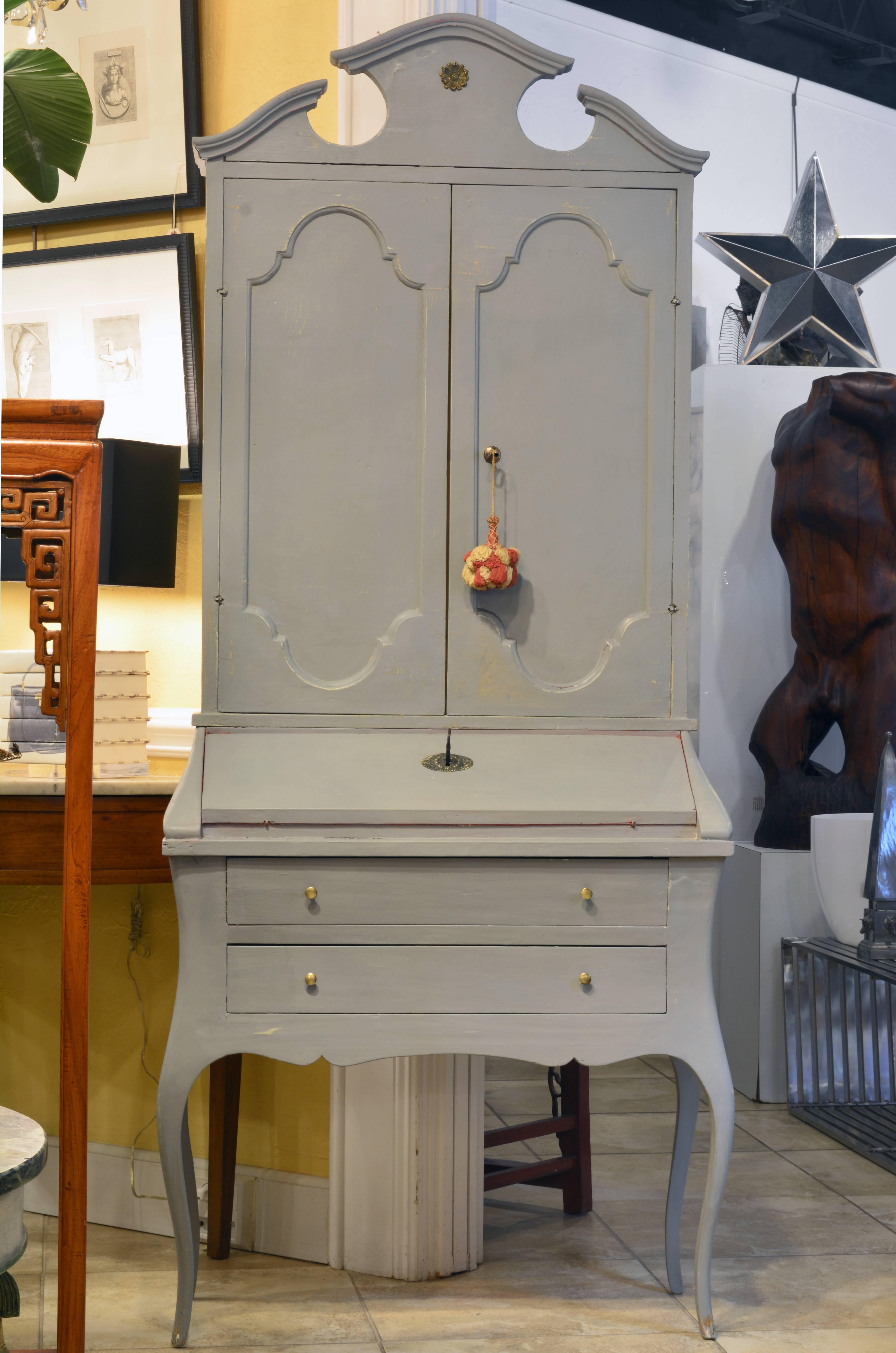 This fetching two part grey painted secretary desk features an upper cabinet crowned by shaped pediments above two-panel doors opening up to a shelved and paper lined interior. The lower part offers a slant front opening up to a Venetian red