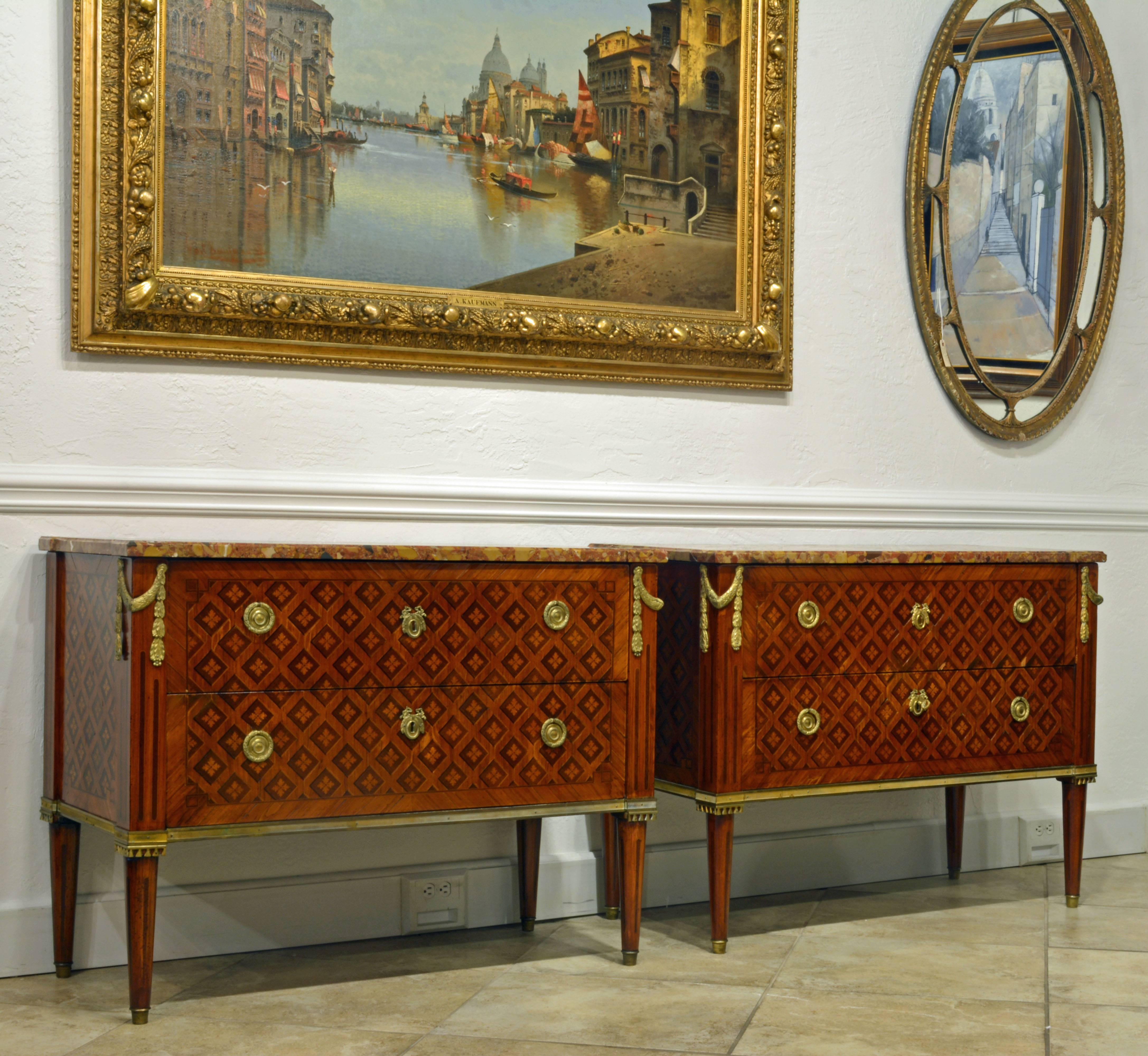These elegant commodes of the Louis XVI era feature shaped variegated marble tops above two long drawers sans traverse, the bottom drawer with lock, the fronts and sides with elegant diagonal pattern tulipwood and kingwood marquetry, the corners