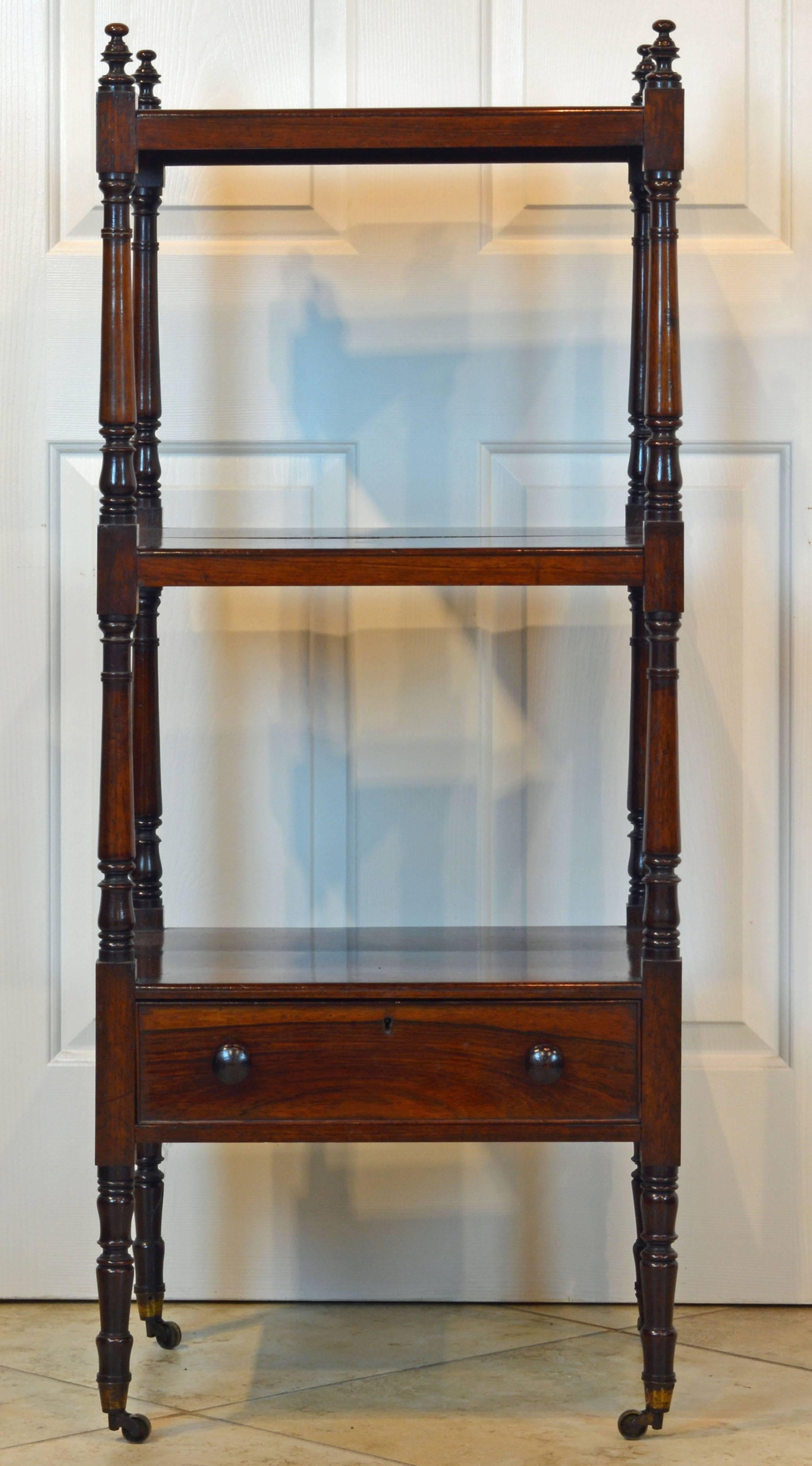 This late Georgian rosewood étagère features three shelves raised on elegantly turned and carved supports surmounted by finials and ending in brass casters. Under the lower shelf a long drawer with turned knobs.