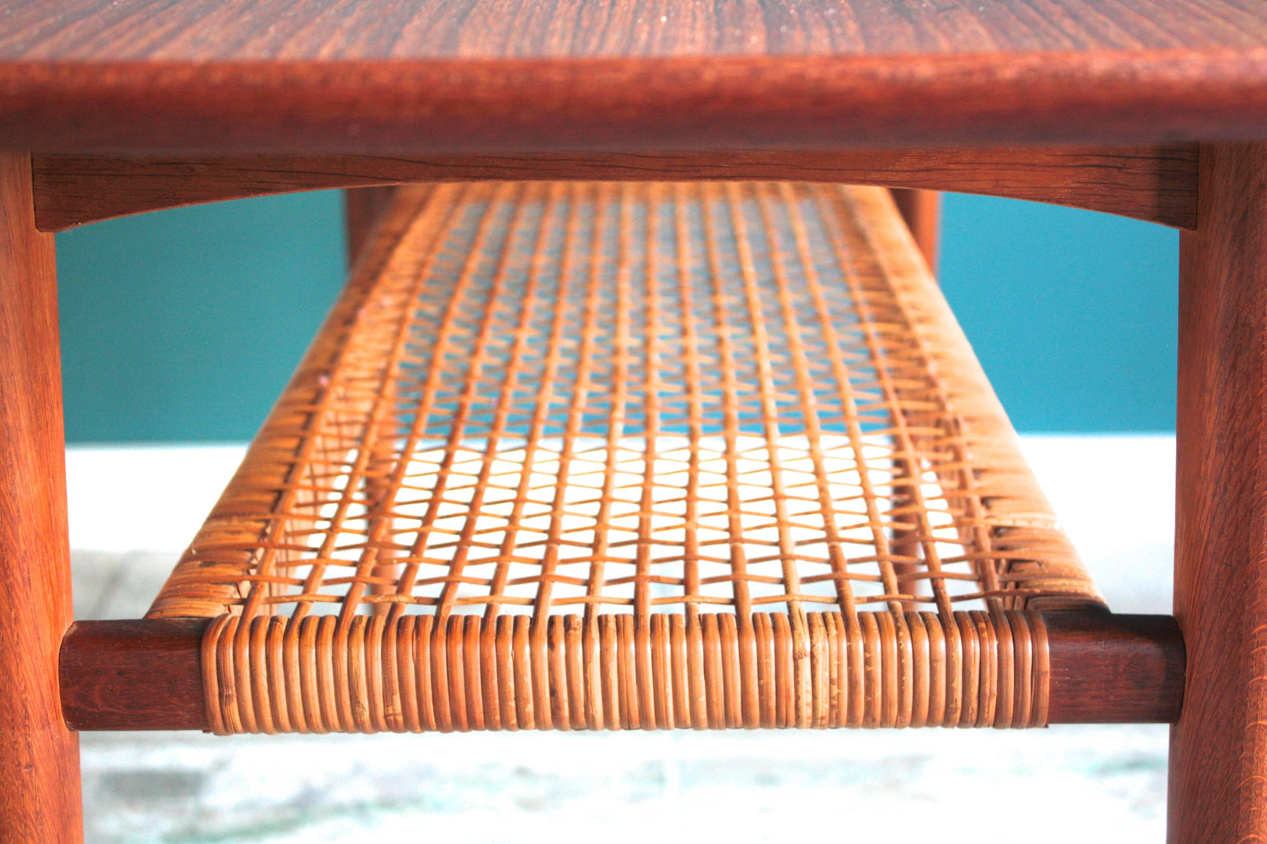 This teak coffee table with magazine rack was designed during the 1950s, and manufactured in Denmark. Made from teak, it features a rattan magazine shelf beneath the tabletop. The edges of the tabletop are rounded. In very good vintage condition