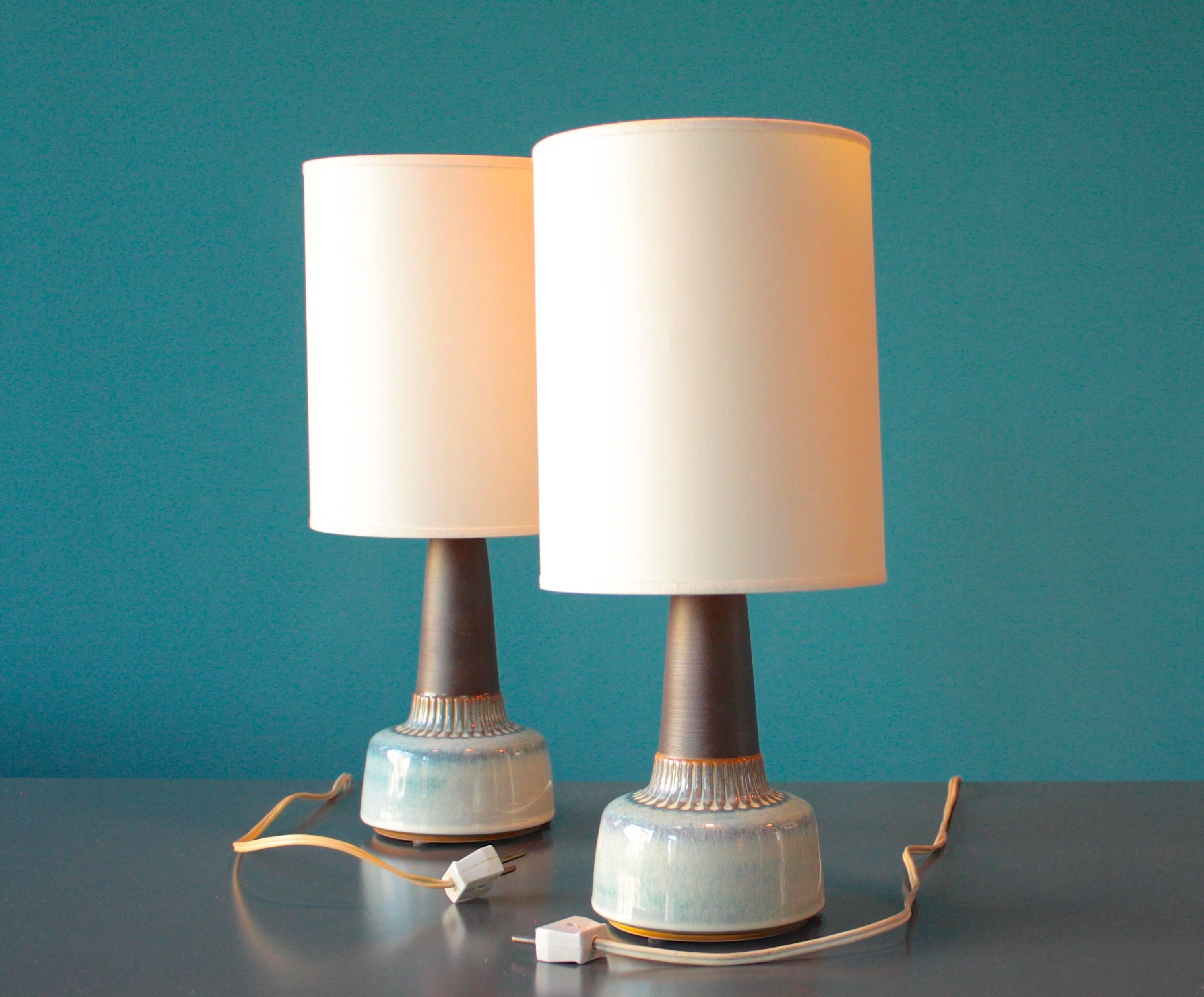 This pair of Mid-Century table lamps was manufactured in Denmark by Soholm, during the 1960s. The frames are made from porcelain and the shades from white fabric. One of the lamps is two centimeters smaller than the other one. This set remains in a