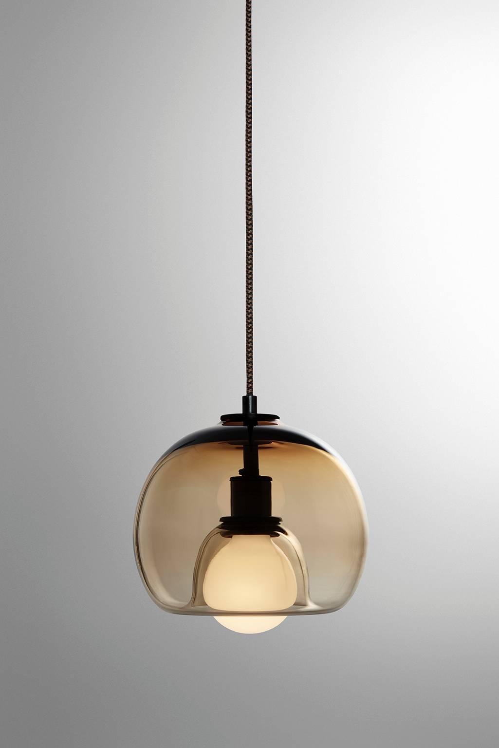 A handblown orb gracefully inverted to house a globe bulb, like a pearl encased in a glass oyster. The Eres bronze has an 8” diameter with a 4' black and brown houndstooth fabric-covered cord and a 4.75” diameter black porcelain canopy. Houses one