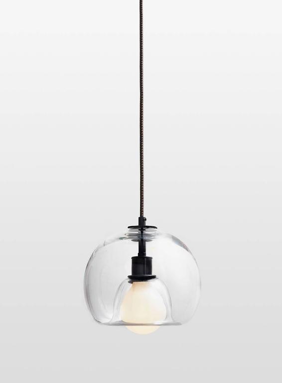 A handblown orb gracefully inverted to house a globe bulb, like a pearl encased in a glass oyster. The Eres clear has an 8” diameter with a 4' black and brown houndstooth fabric-covered cord and a 4.75” diameter black porcelain canopy. Houses one