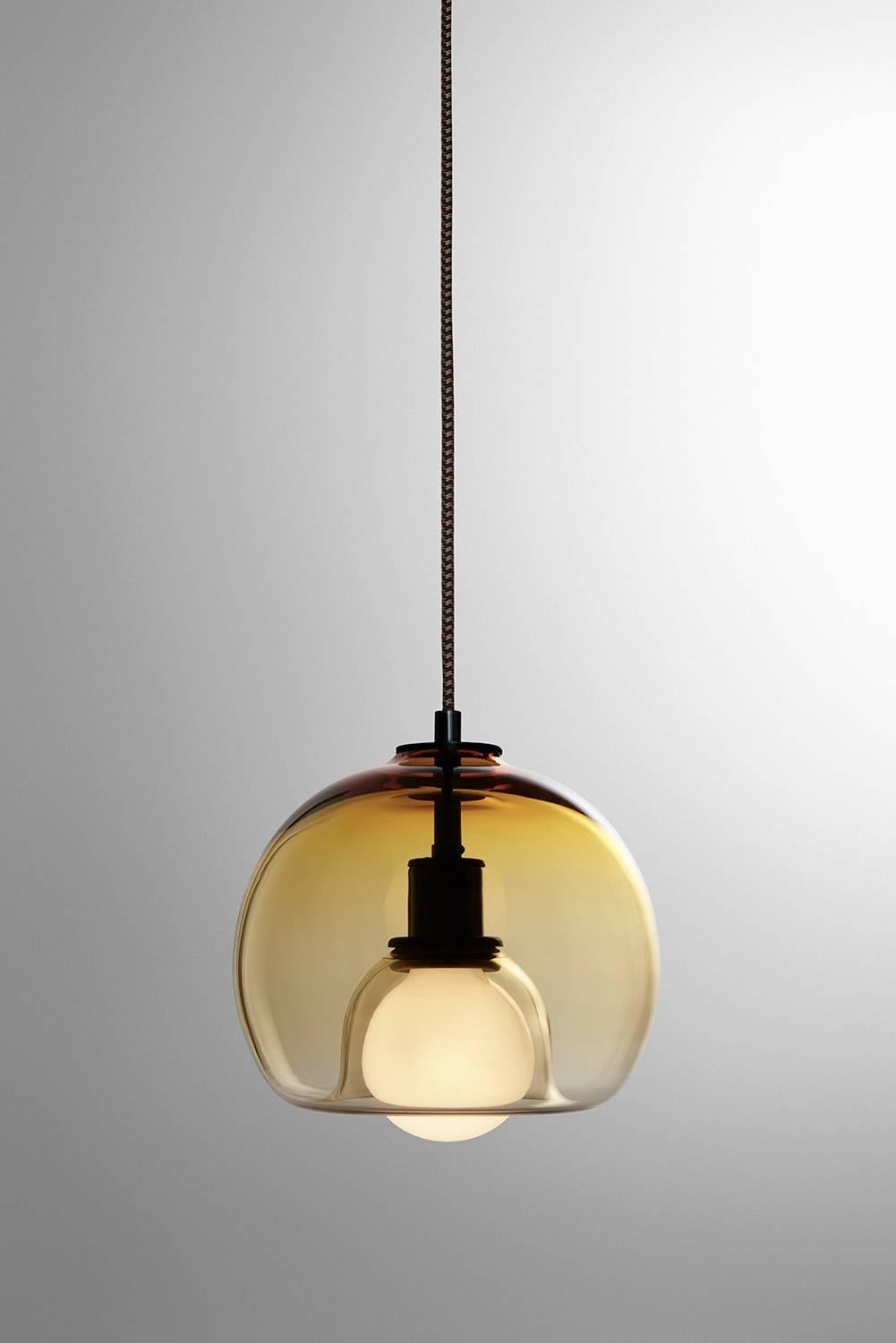A handblown orb gracefully inverted to house a globe bulb, like a pearl encased in a glass oyster. The Eres Gold has an 8” diameter with a 4' black and brown houndstooth fabric-covered cord and a 4.75” diameter black porcelain canopy. Houses one