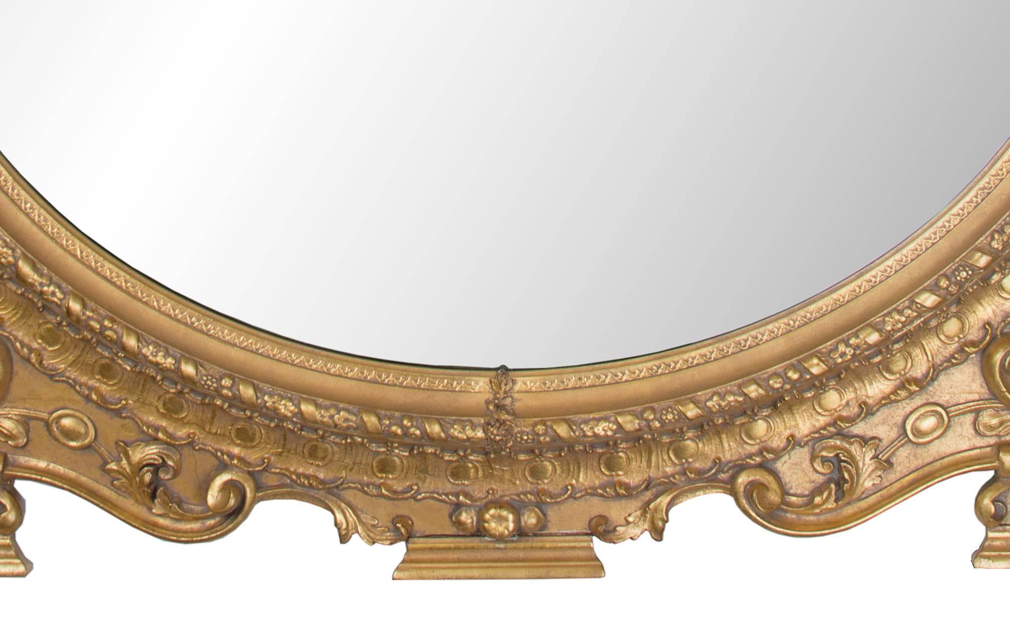 19th century carved giltwood and composition overmantle mirror. Imposing central crest with foliate and scrolled decoration over the oval mirror plate framed with egg and dart decoration and floral rope design. The over-all mirror rests on three
