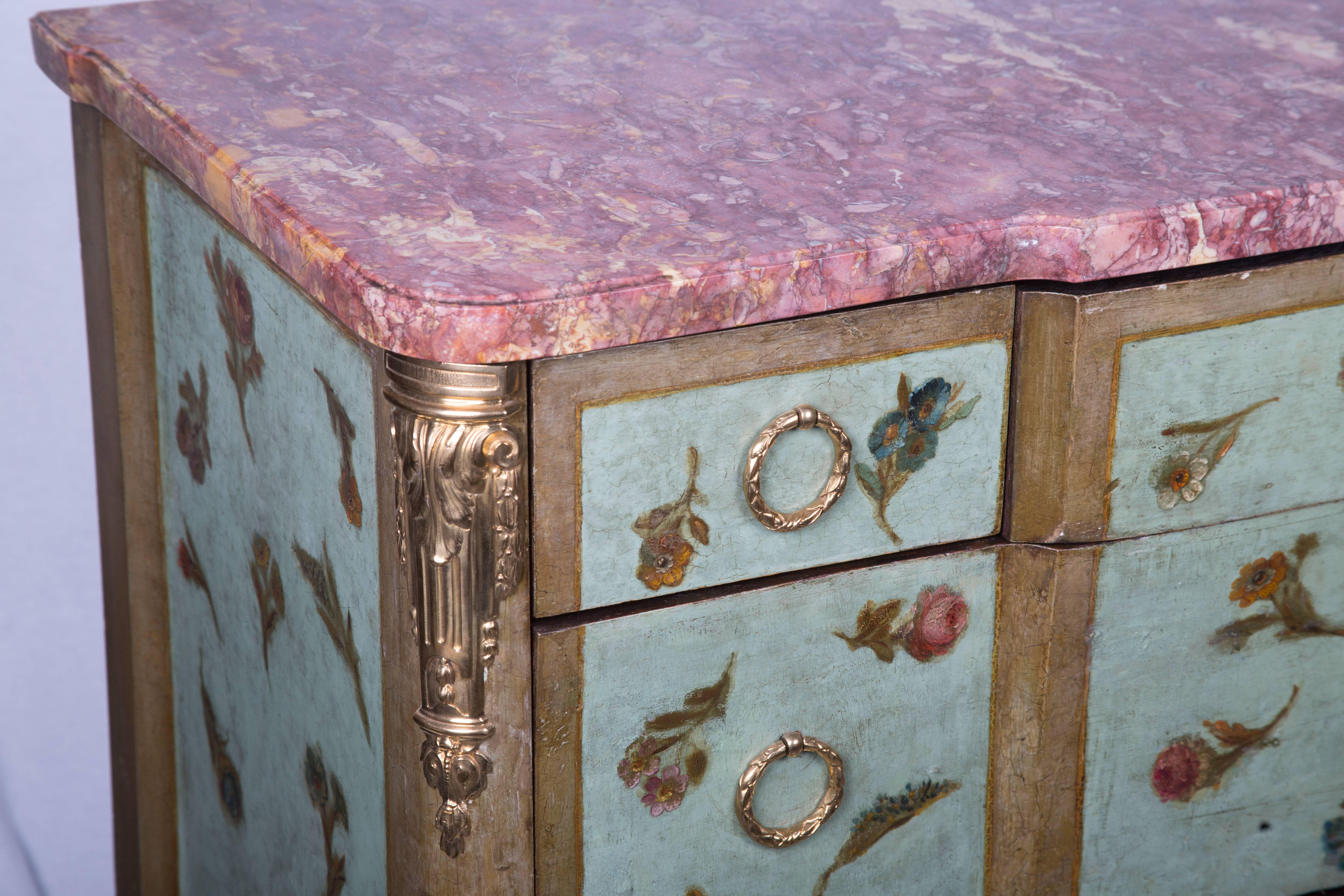 A softly decorated Louis XV/XVI transitional style commode, painted over-all with flowers on a light blue/green background. The variegated marble breakfront top is over center drawer flanked by two short drawers and two long graduated drawers