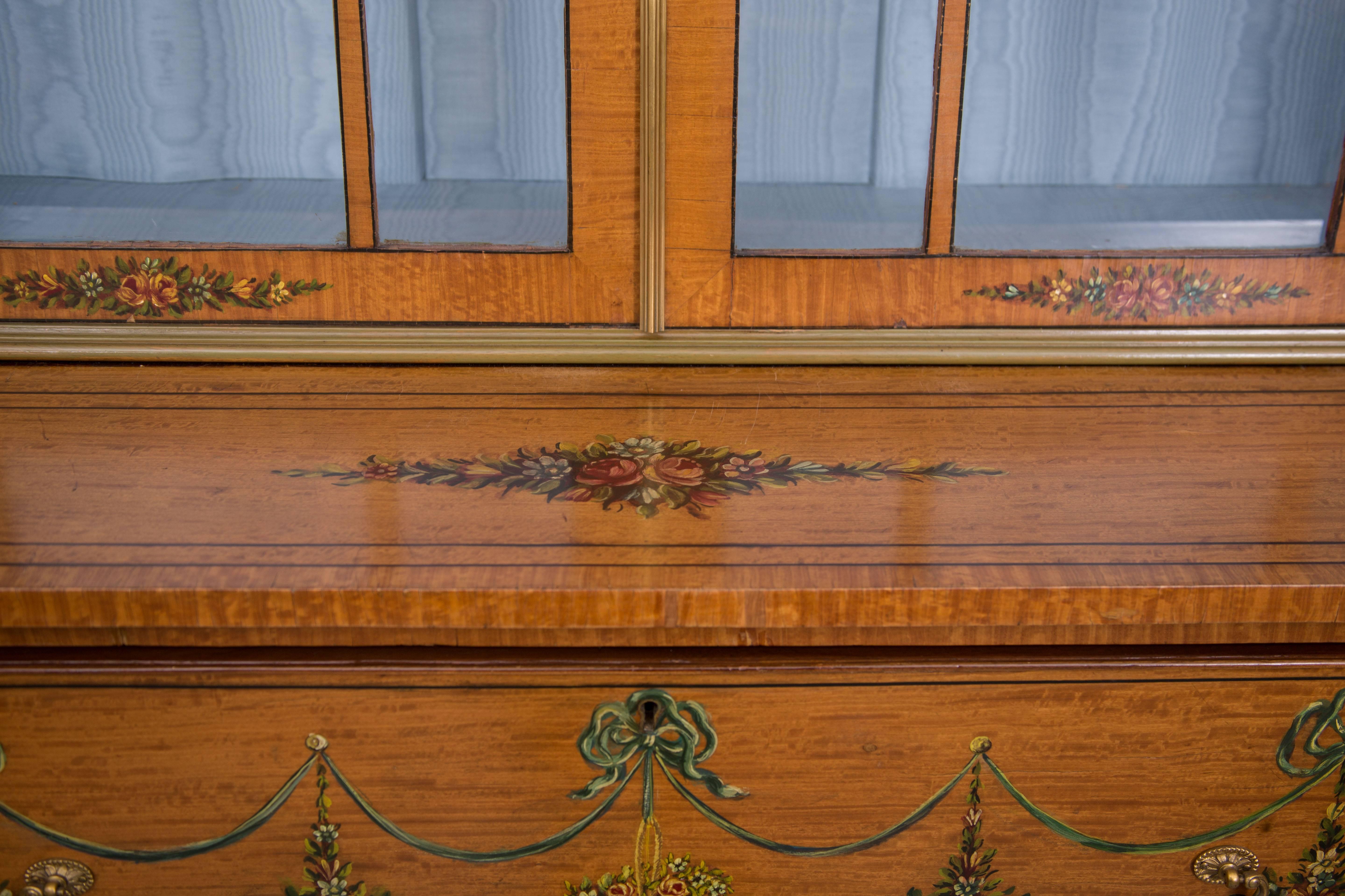 This is a fine example of an English Adams style painted satinwood secretary painted overall with delicate flowers and swags. The top section has a prominent cornice over two glazed astragal doors with arched mullions, opening to reveal sky blue