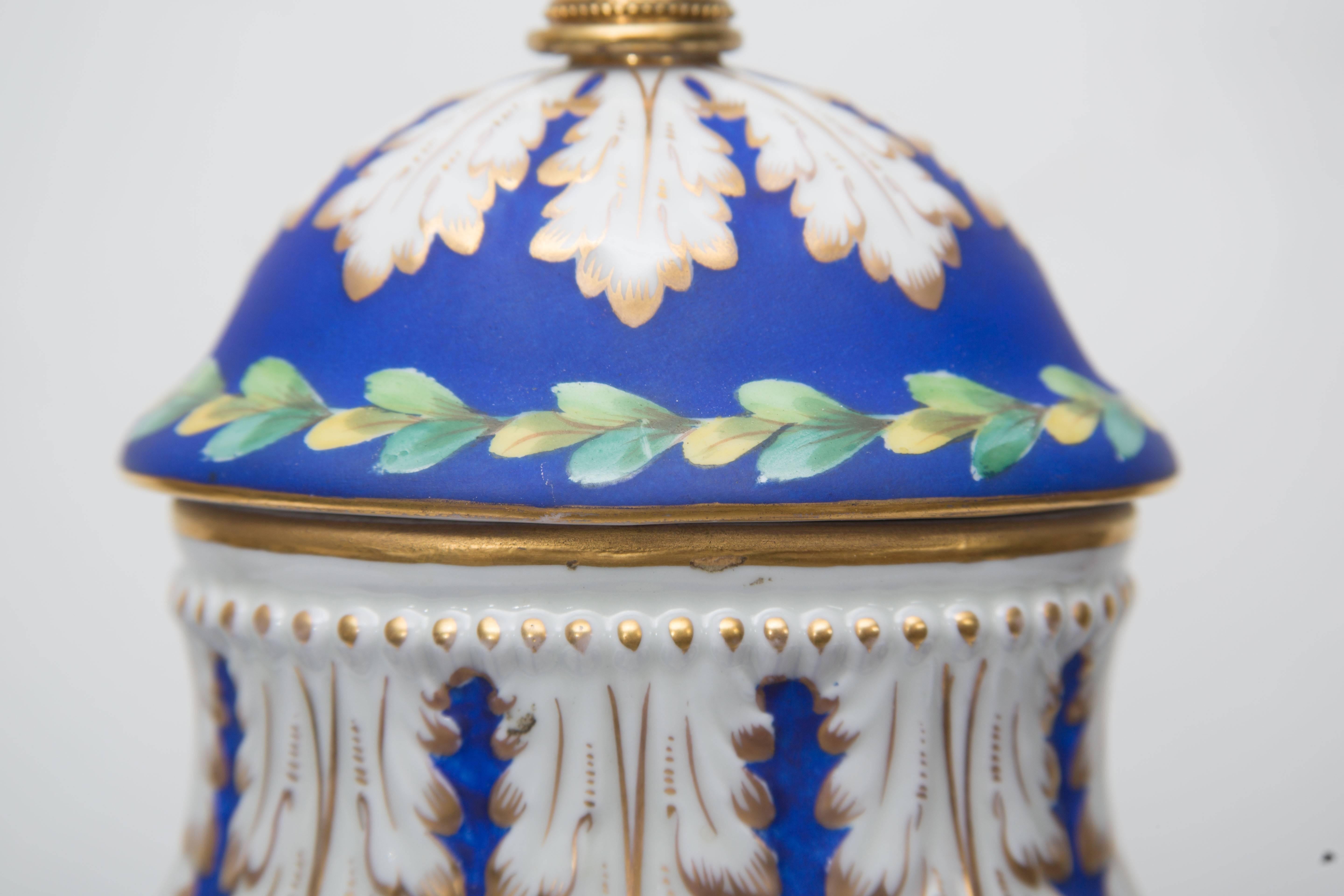 This is an elegant French glazed porcelain and bisque urn converted to a lamp. The oval-form vase with sphinx busts has a bisque section with painted flowers in a decorated basket on a blue background juxtaposed to white glazed and gilt heightened