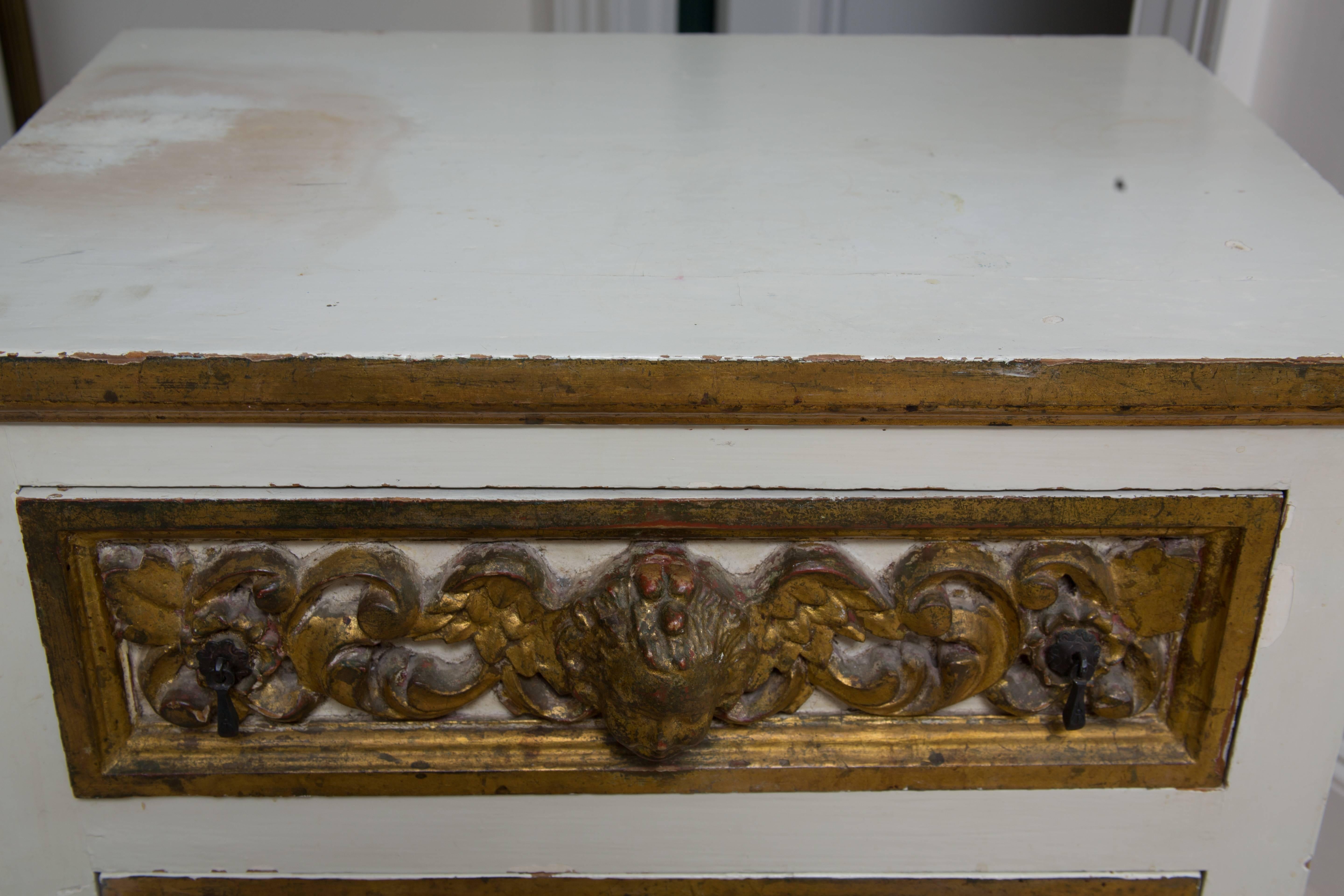 This lovely pair of Italian white painted and parcel-gilt three-drawer chests have drawer faces heavily carved with a central winged mask flanked by scrolls. The sides are designed with raised gilt blocking framed by antique mirror. 19th century.