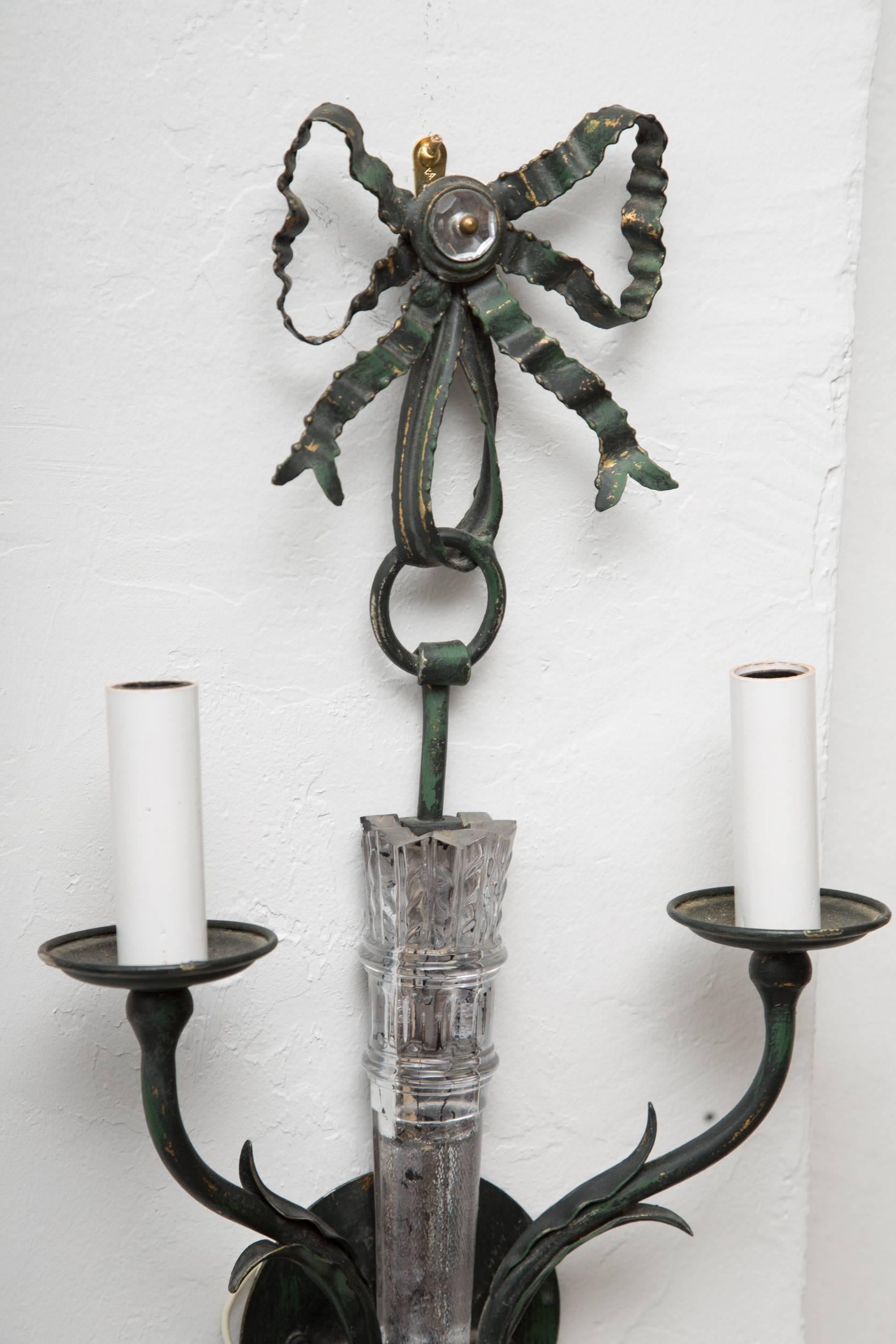 This pair of painted metal sconces has a central body in the form of a glass quiver, appearing to be suspended from a ring attached to a bow, and flanked by two out-scrolled electrified candle arms. 