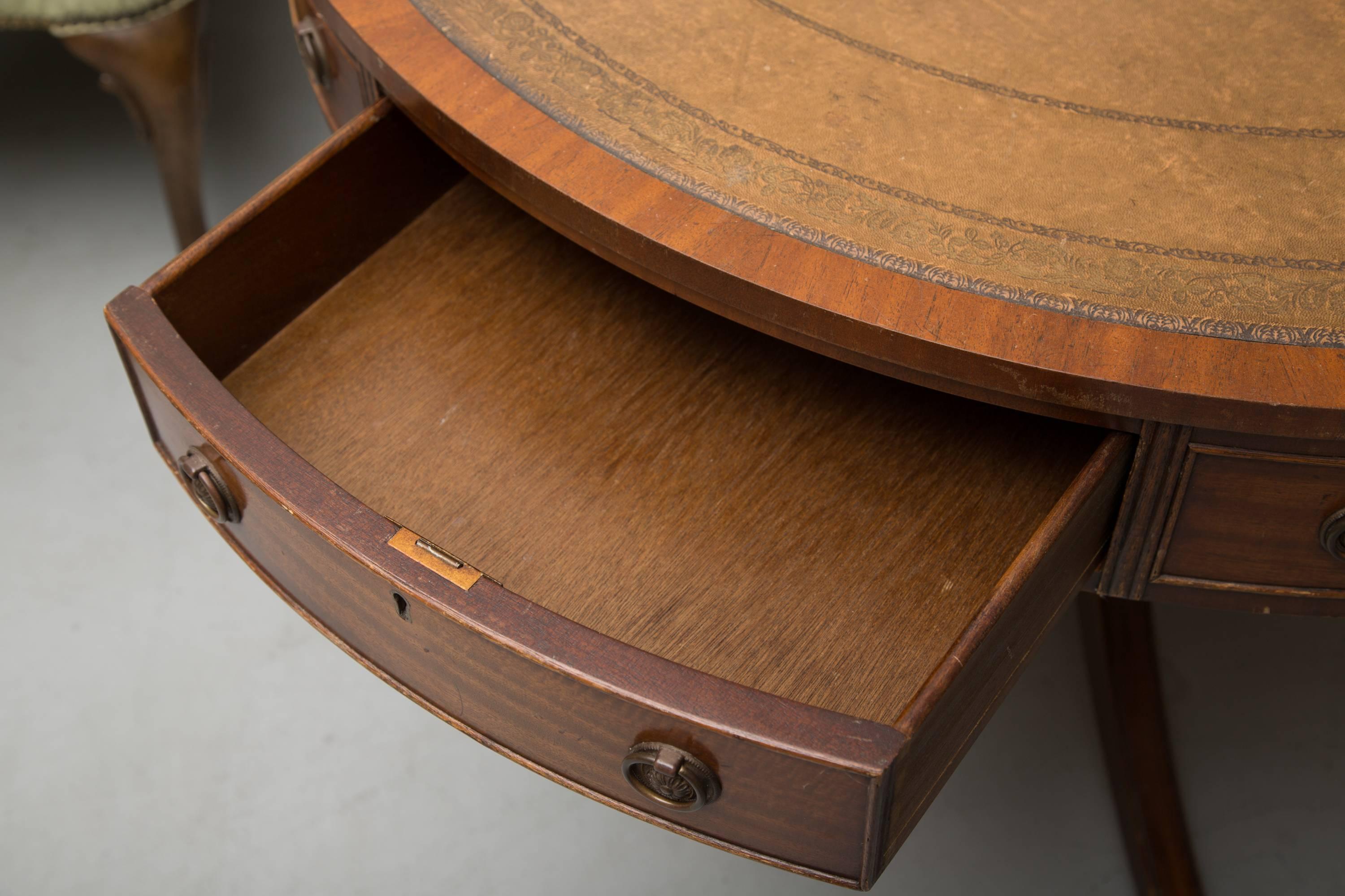 This 20th century mahogany bench-made circular drum table has a tanleather inset with gilt tooling framed by mahogany crossbanding. The wide frieze has a combination of faux and functional drawers accented with Fine beading. The circular structure