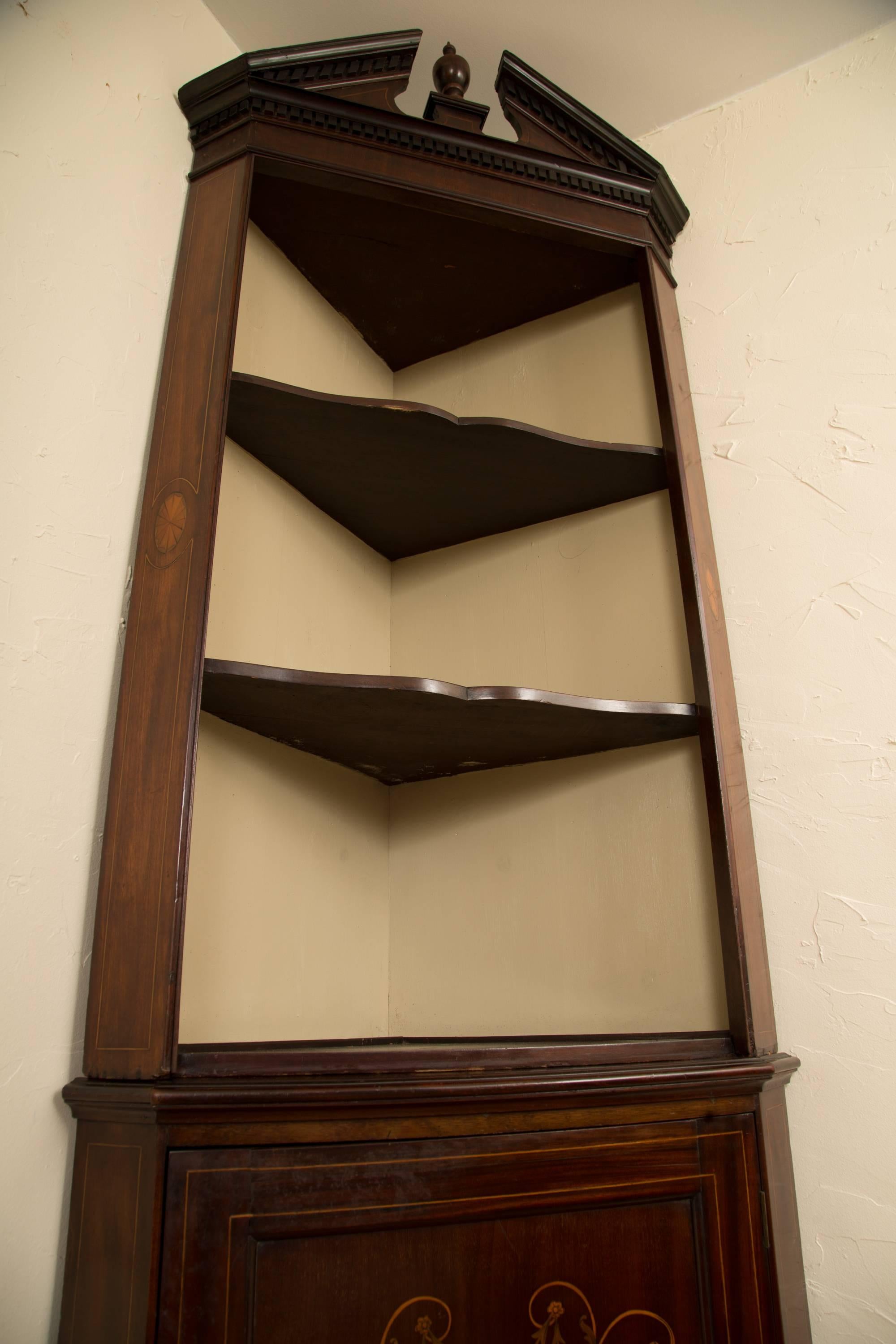 This is a tall, stately English Edwardian corner cabinet in the Georgian style. The top has a broken swan neck pediment centred by an urn finial and a frieze with dentil mounding, above an open cavity revealing three shaped shelves, flanked by