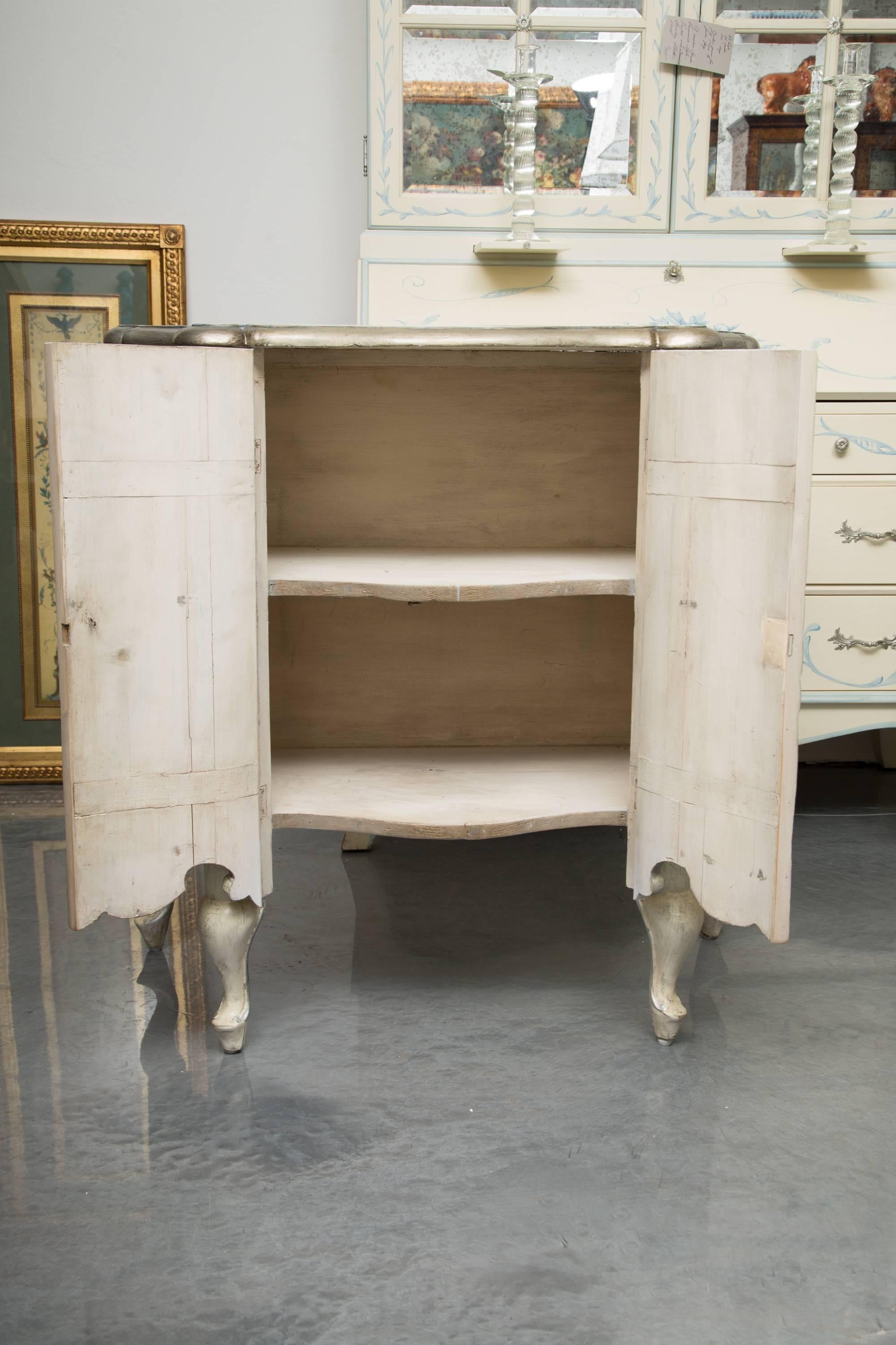This is a sophisticated pair of blue-washed and cream-painted serpentine two-door cabinets with parcel silver highlights. The top with beveled edge is over a pair of paneled doors and inverted serpentine sides, conforming to the top. Each cabinet is