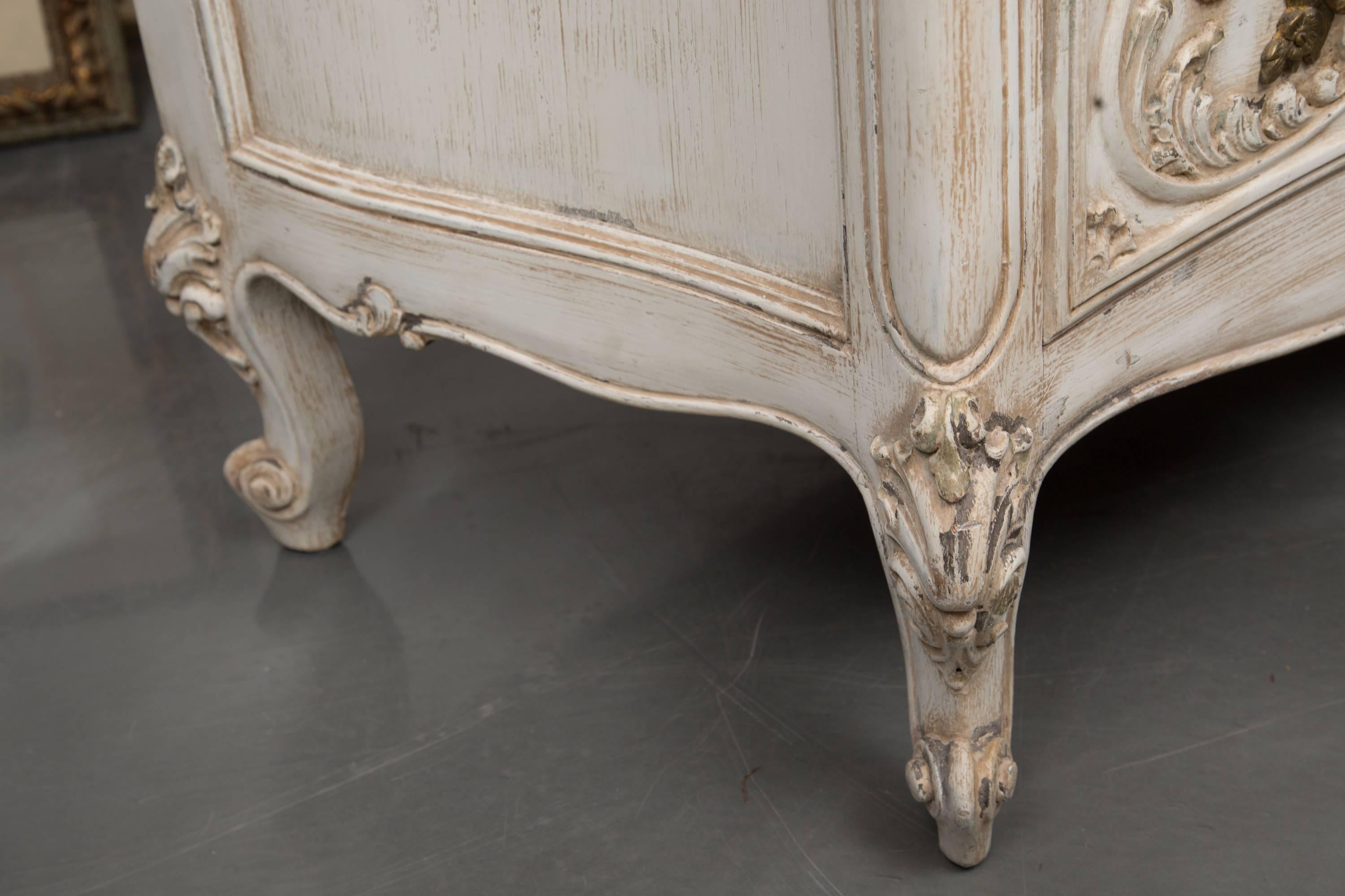 This is an artfully carved cream-painted commode.  The mottled gray marble top with prominent ogee edge is over three long drawers above a carved apron and raised on carved cabriole legs, 20th century