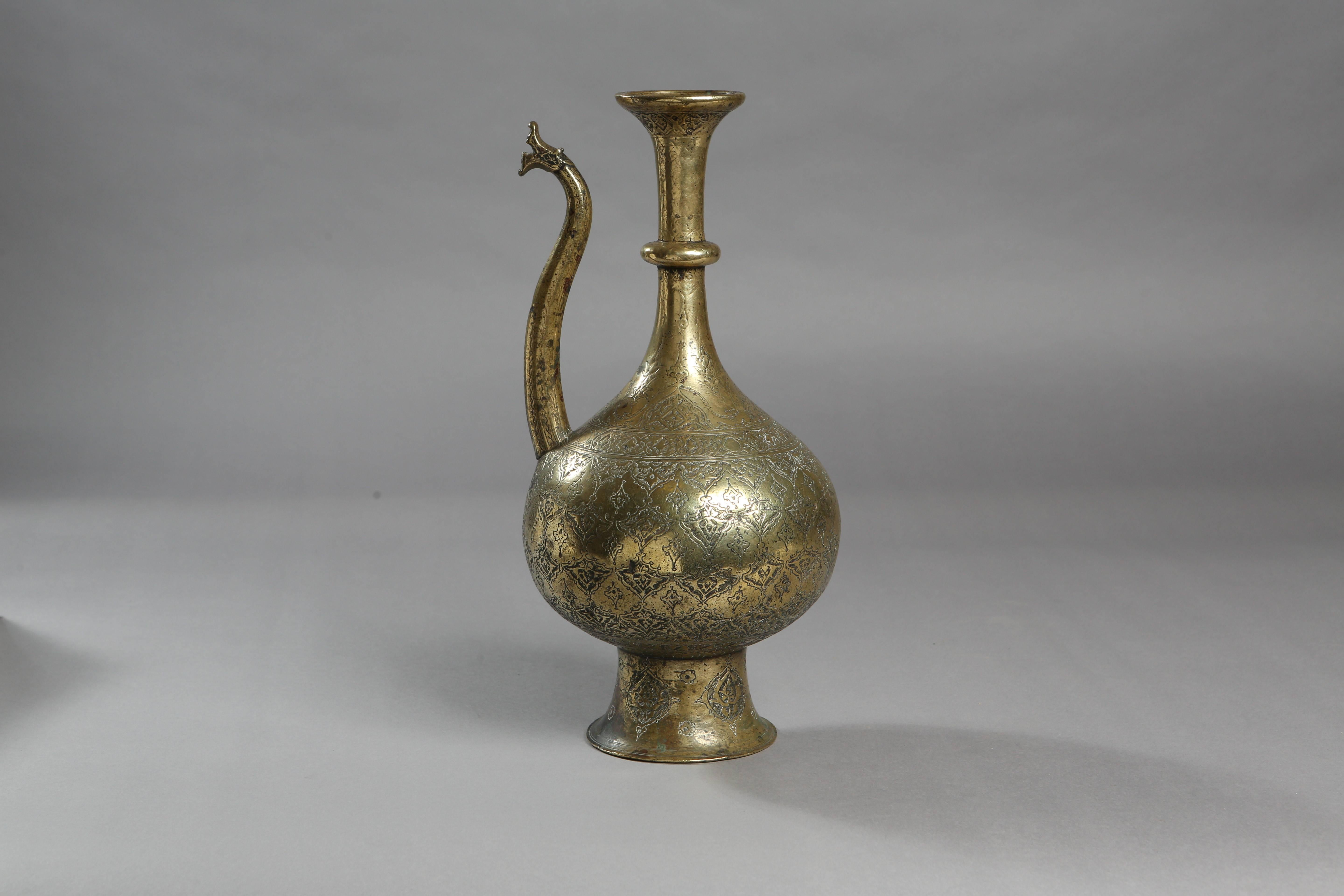 This 16th century Safavid tinned brass ewer is made of a pyriform shaped body engraved on the body, neck and foot with palmettes. The spout is of a serpentine shape with a dragon-head finial and it is in good condition, with normal wear for its