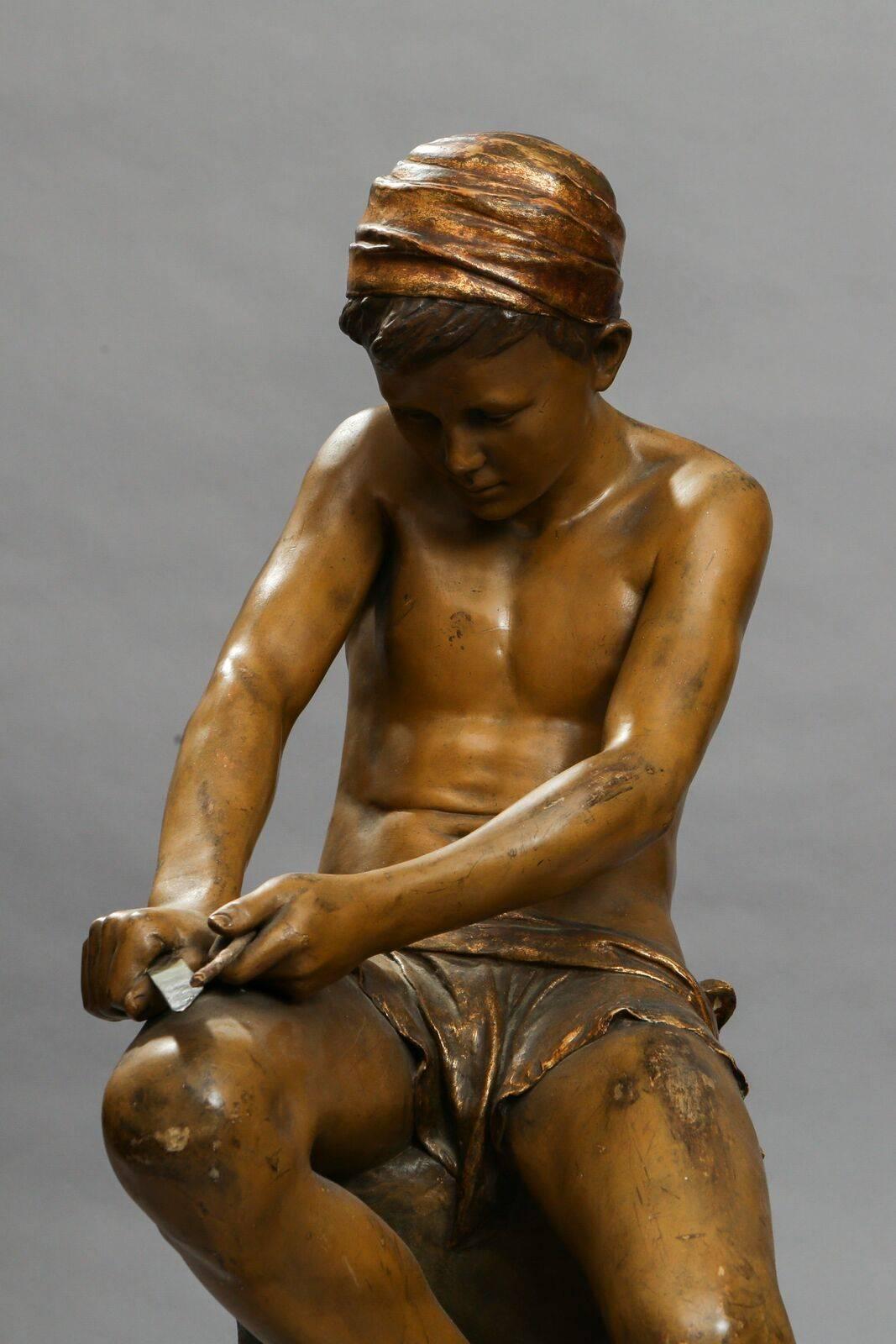 A polychrome terracotta figure of a young seated boy carving. He is holding a metal knife in one hand and is sharpening/carving a stick in the right hand. The figure is signed and marked on the back of the base with the numbers '1067/58/24' and a
