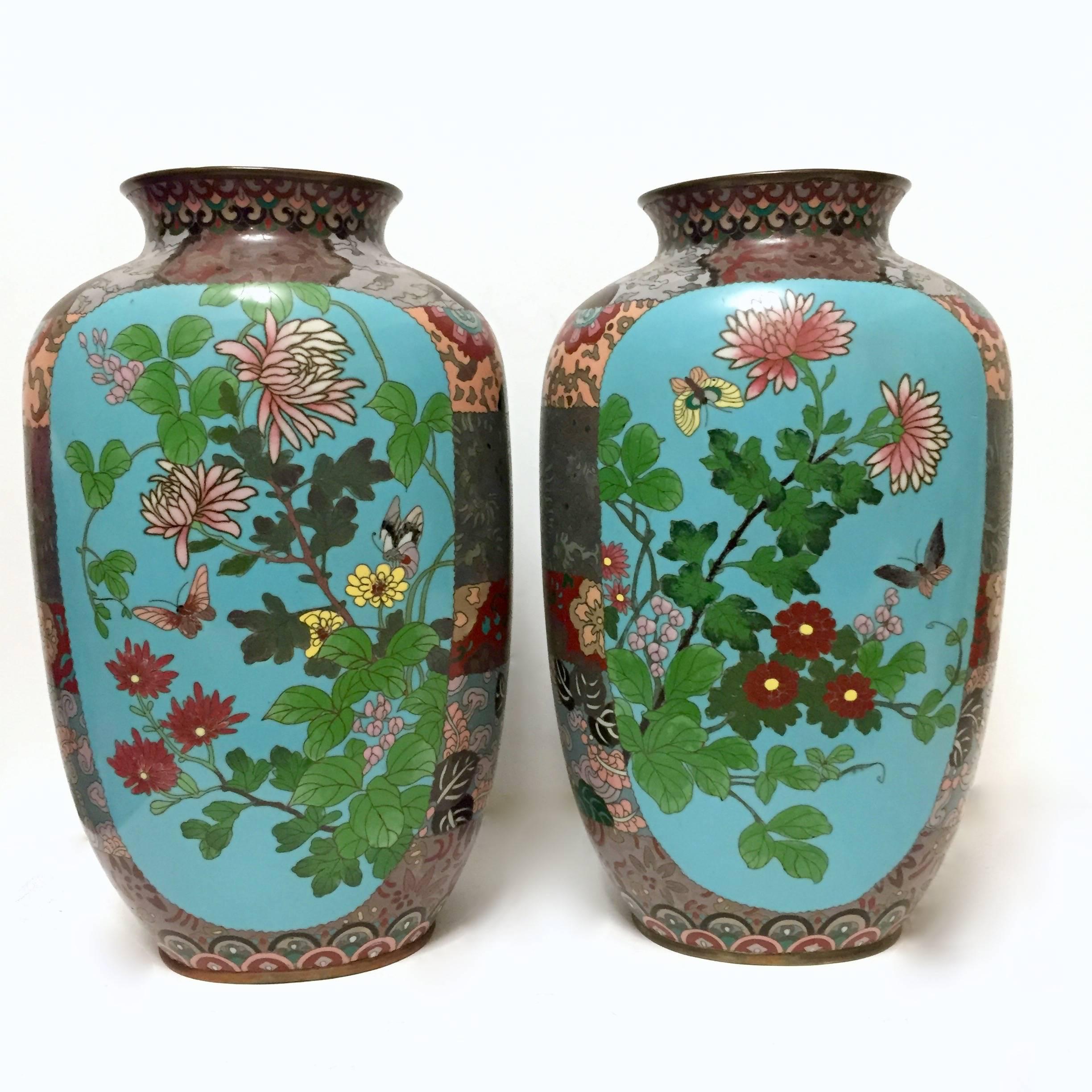 A pair of Japanese cloisonné enamel vases of slightly compressed tapering form. They are heavily ornamented on the neck, shoulders and sides with various motifs and contain central panels of a light blue background that each feature: Butterflies,