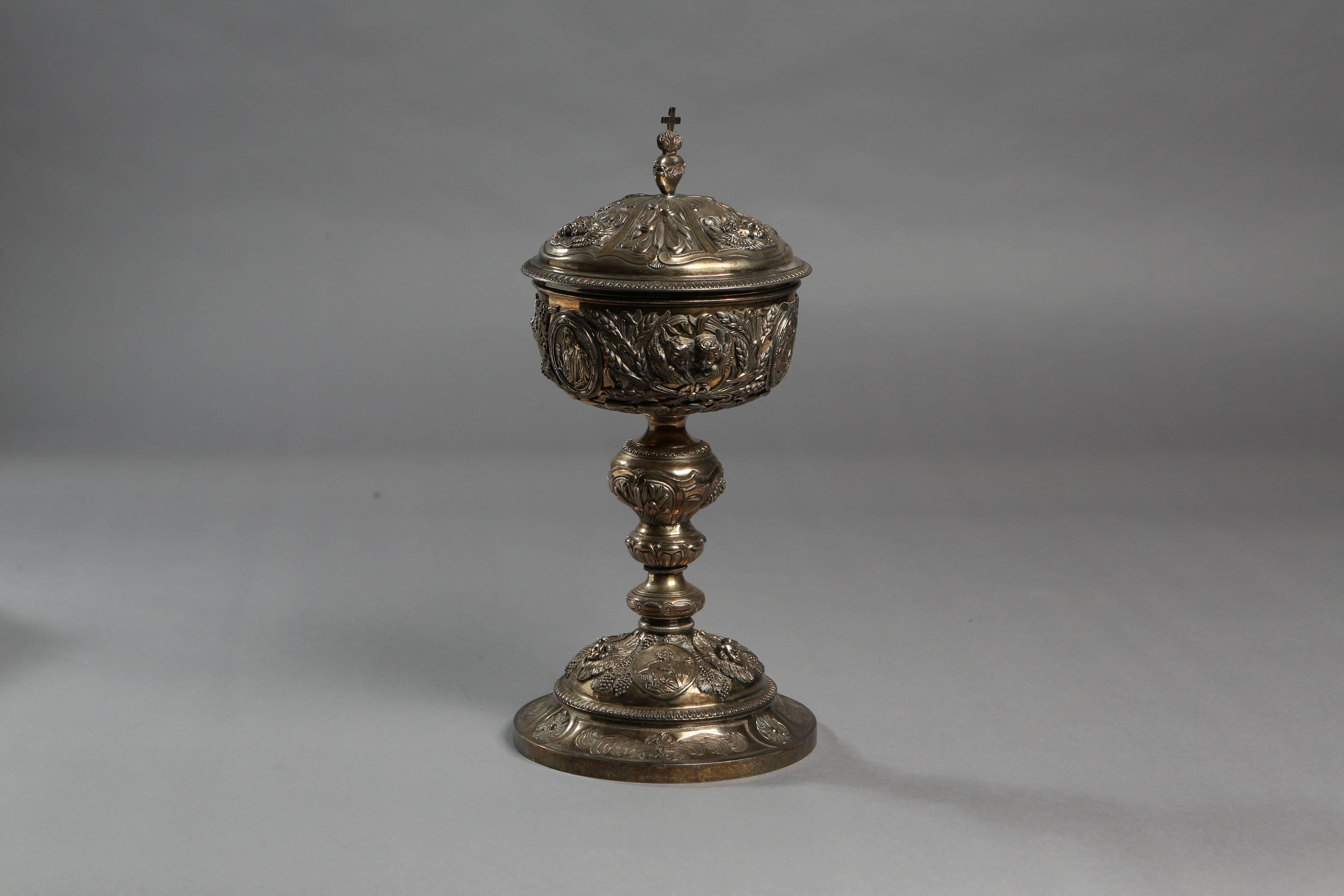 A silver gilt ciborium that is decorated with medallions of the saints on the base and the cup of the chalice. The bottom features depictions of Jesus wearing a crown of thorns and holding the pastoral staff, the ascending Virgin Mary, and Saint