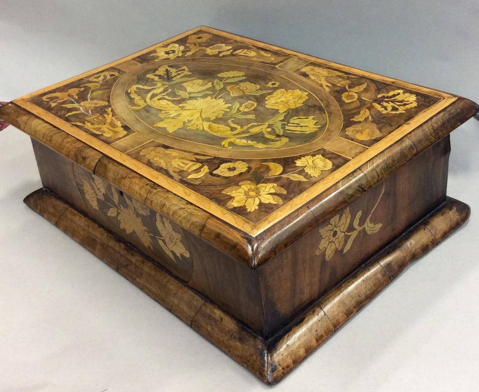A wooden box made of thick panels decorated in floral marquetry. Once stained, the wood shows lighter contrasting designs set against a darker ground. The lid features a rectangle framing an ovular central design with bouquets fitting in the divides